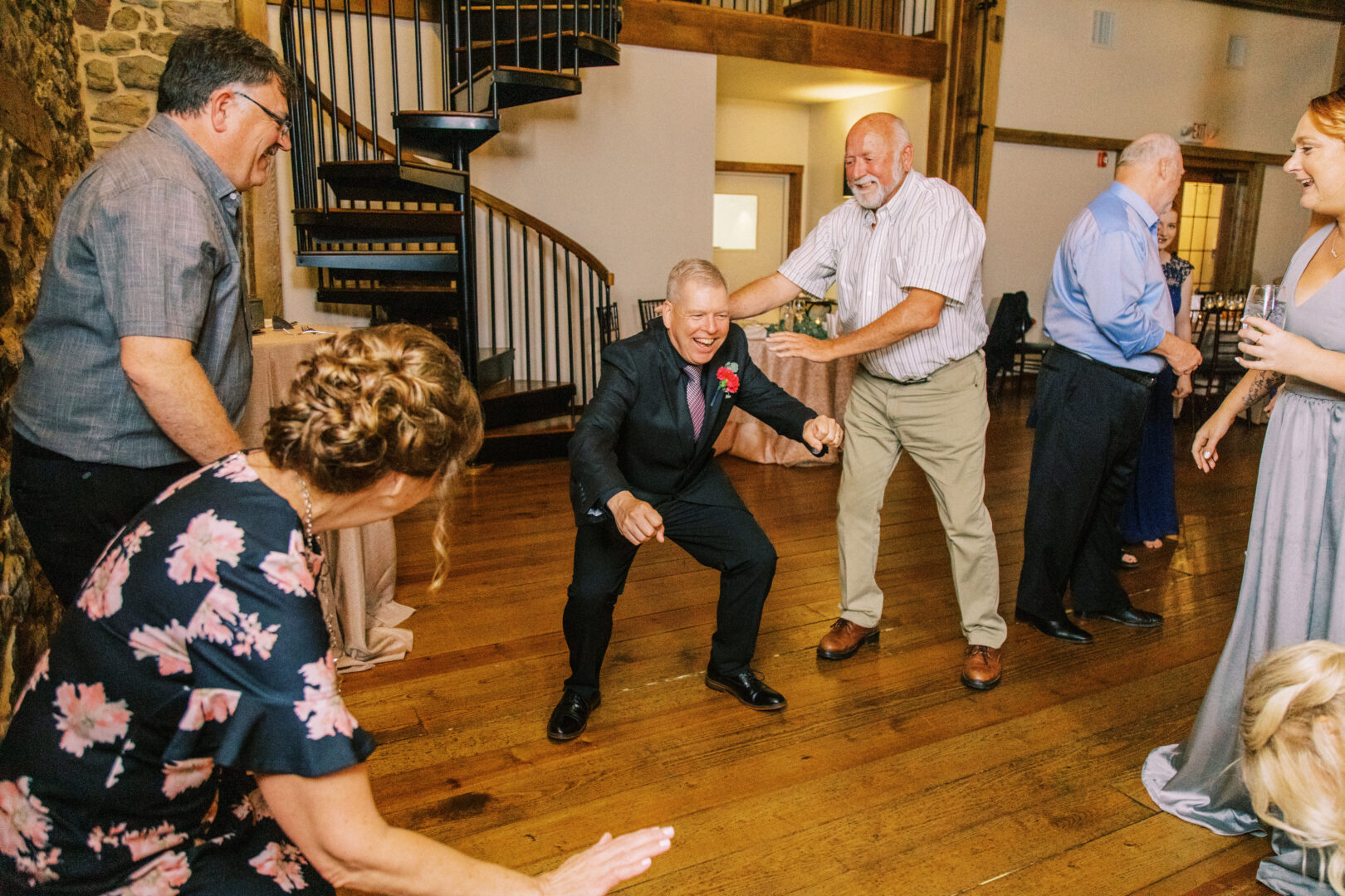 A group of people dances and laughs together on a wooden floor near a spiral staircase, reminiscent of the lively atmosphere at a Harvest View Barn at Hershey Farms wedding during an intimate indoor gathering.