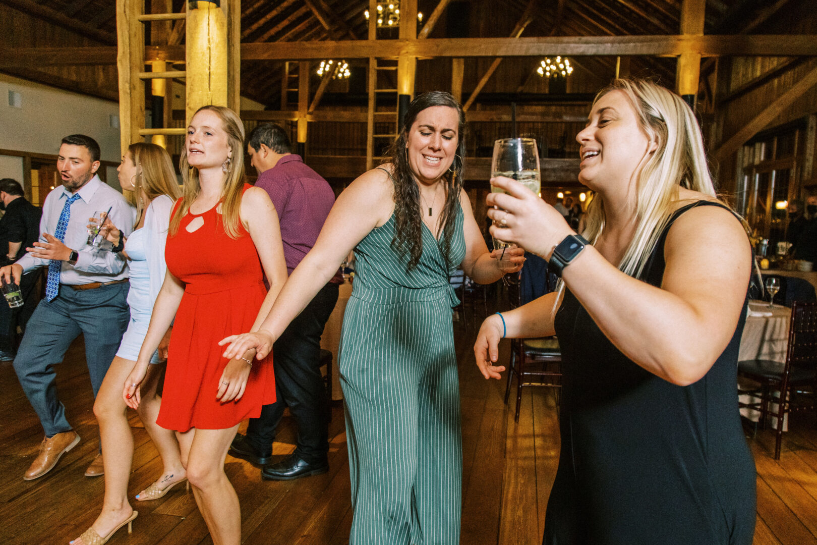 At the Harvest View Barn at Hershey Farms Wedding, people dance and hold drinks in a warmly lit venue adorned with wooden beams and chandeliers.