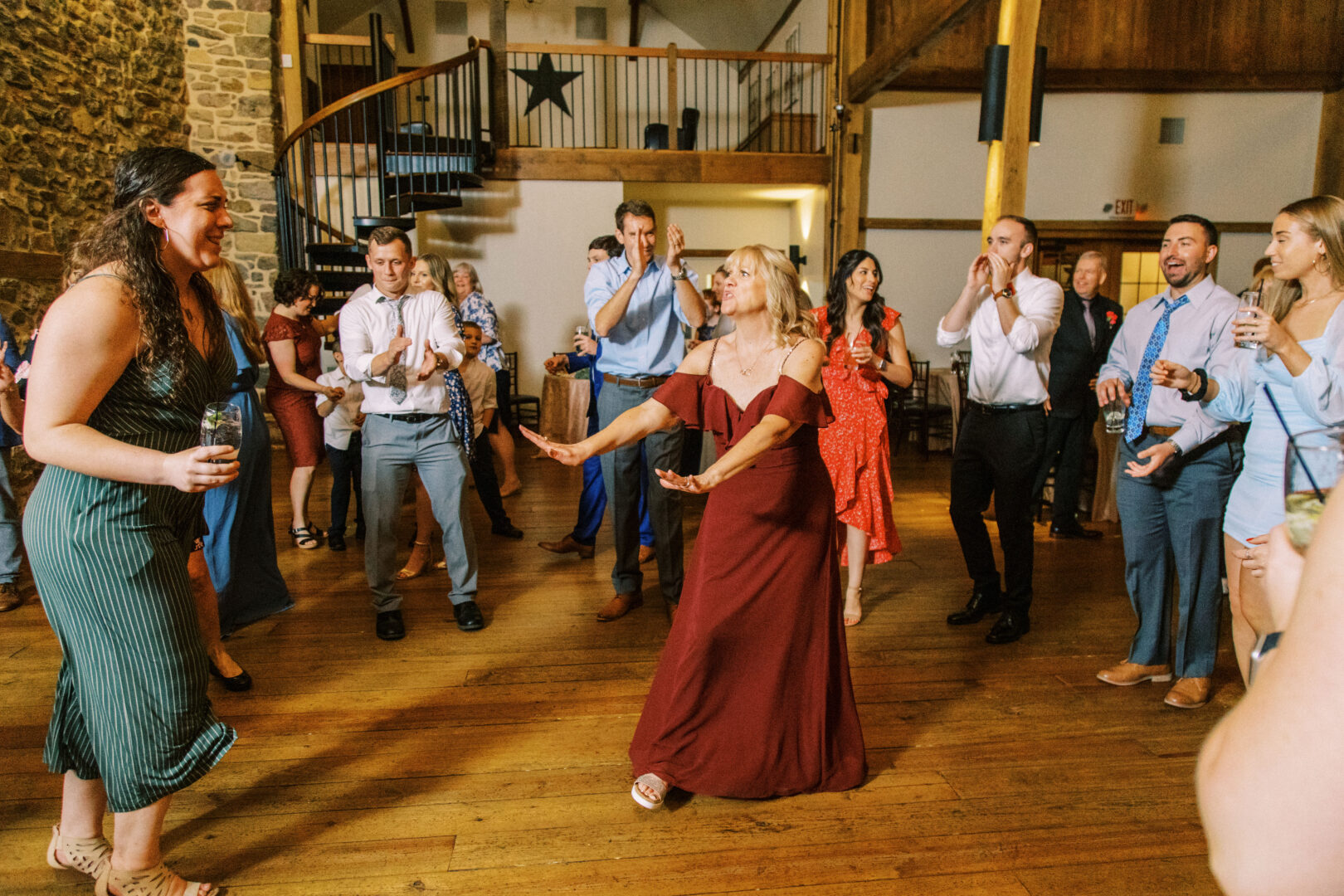 In the rustic charm of Harvest View Barn at Hershey Farms, a group of people are dancing and clapping on wooden floors beneath stone walls. At the center, a woman in a red dress swirls gracefully, embodying the joy of the celebration.