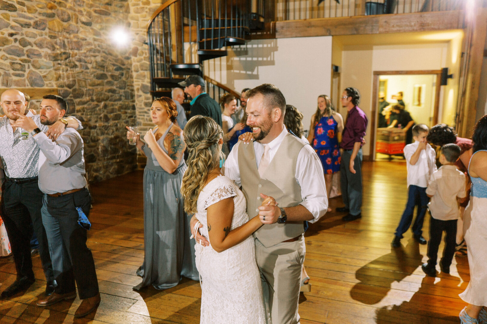 A couple dances in a warmly lit venue with a stone wall at the Harvest View Barn. Several other people are seen socializing and dancing around them, adding to the festive atmosphere of this Hershey Farms wedding.