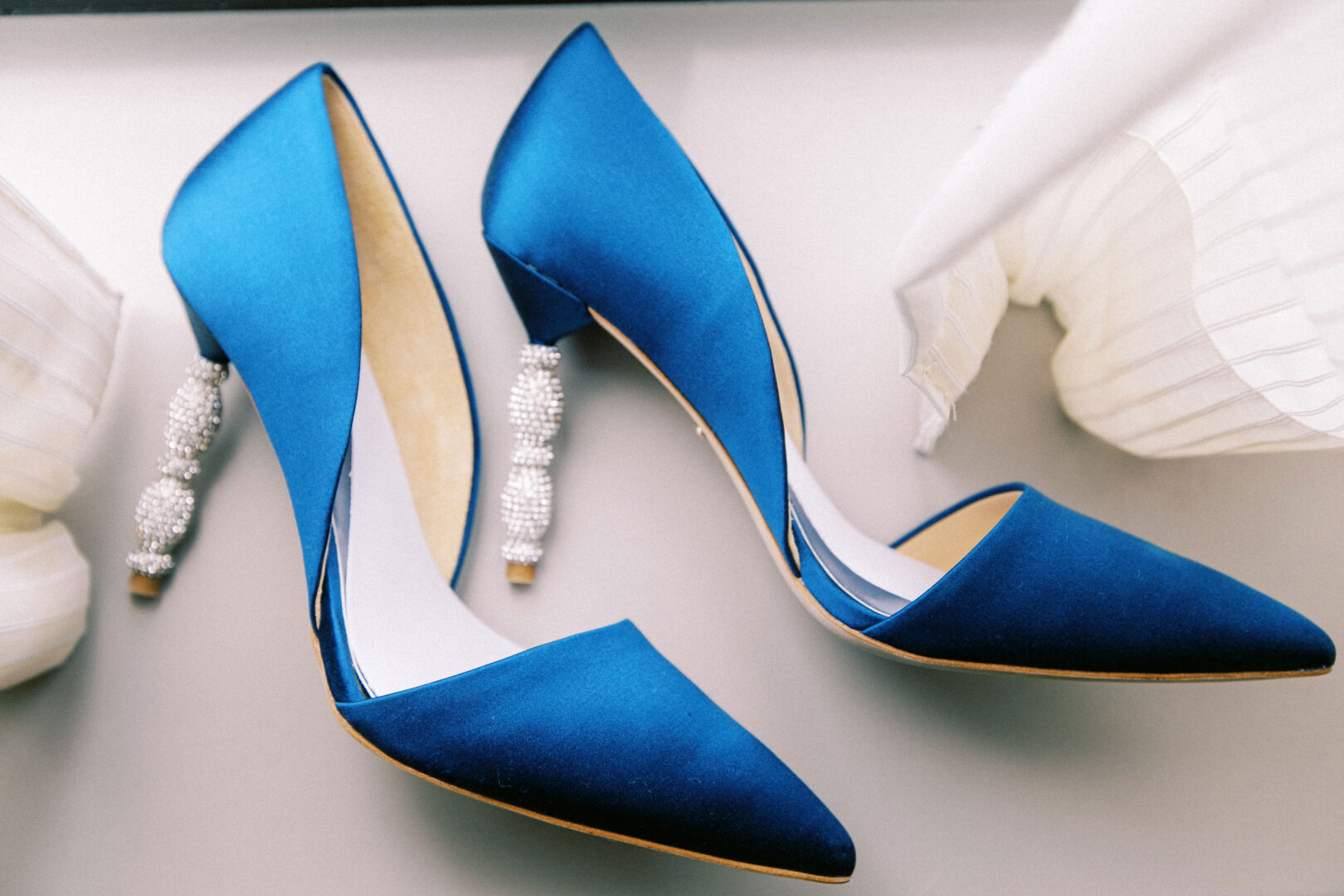 A pair of blue satin high heels with embellished heels are placed on a light surface near white fabric, capturing the elegance of a Mansion on Main Street wedding.
