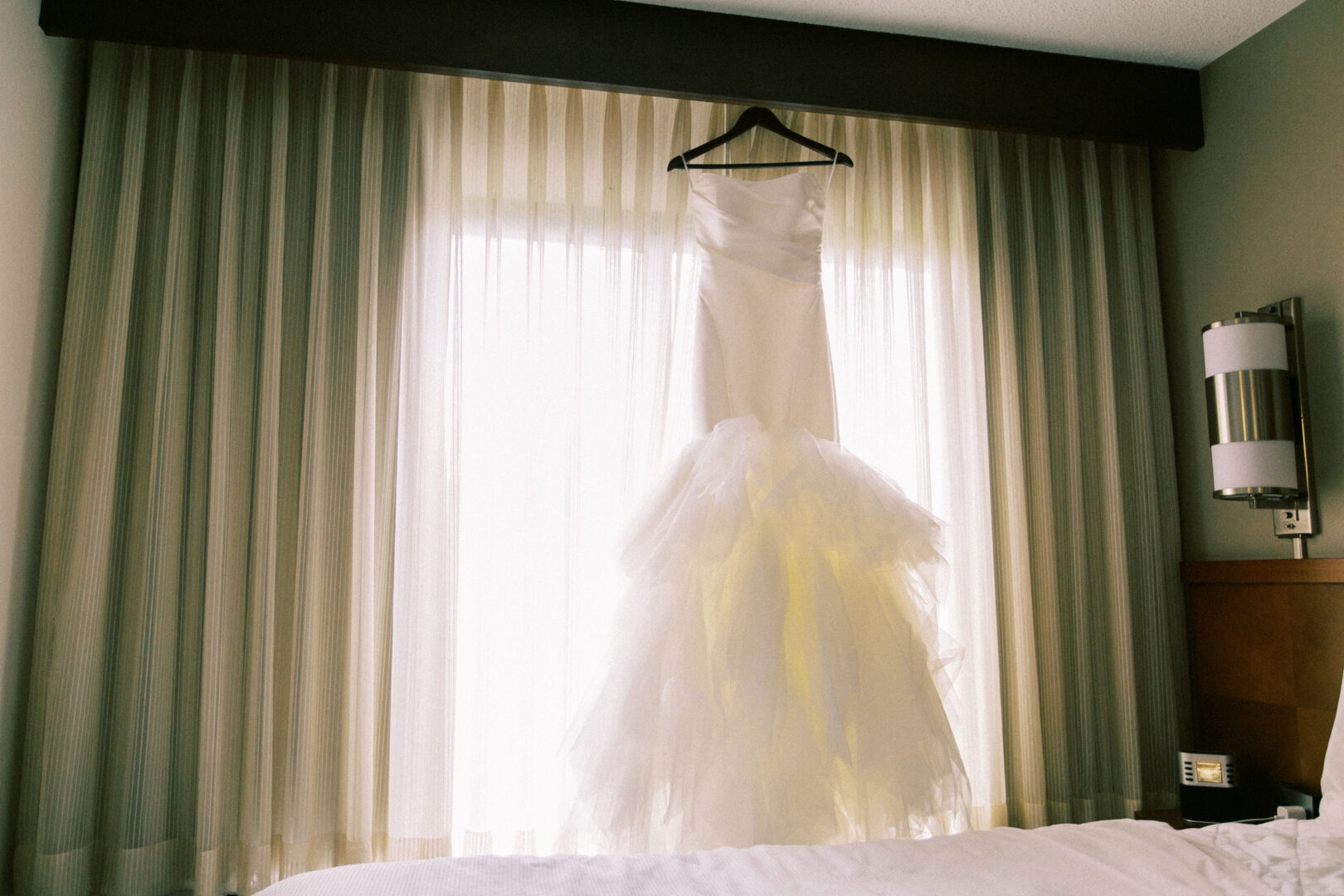 A white wedding dress hangs elegantly on a hanger in front of sheer curtained windows, casting a soft glow, reminiscent of the timeless charm found at a Mansion on Main Street Wedding.