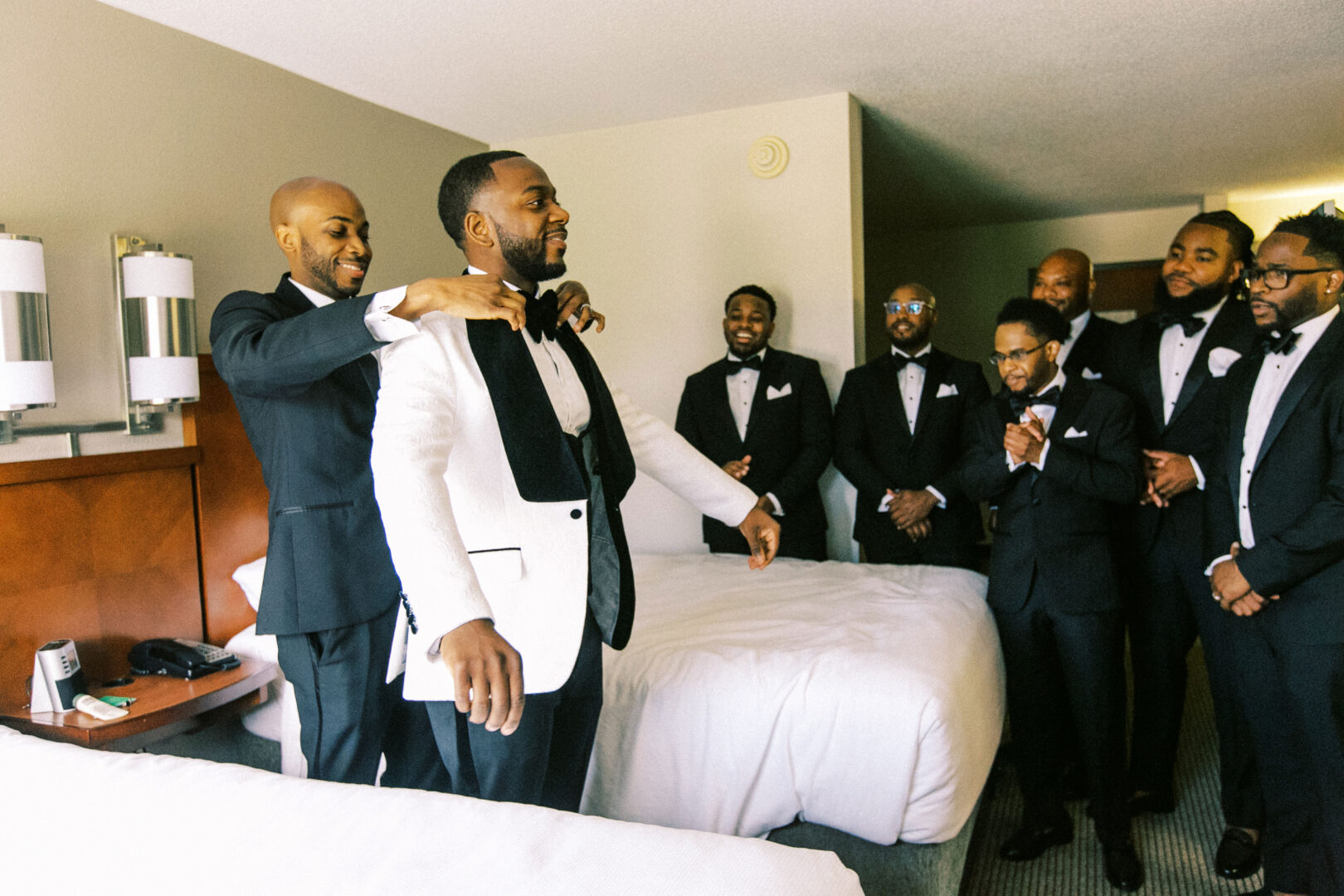 In a refined setting, a group of groomsmen in suits gather at the Mansion on Main Street. One man helps another adjust his white tuxedo jacket as others look on and smile, capturing the essence of this elegant wedding moment.