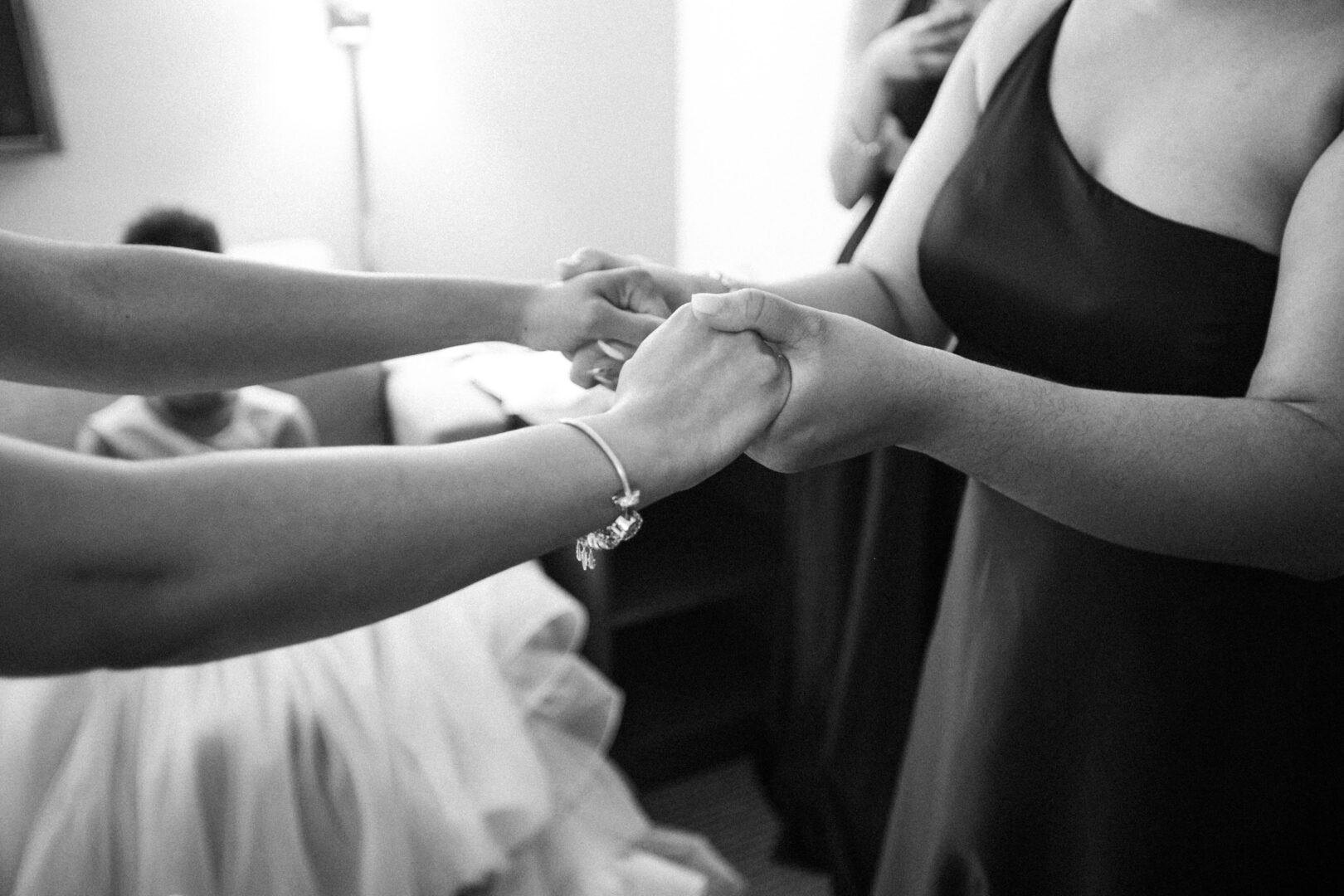 Two people holding hands in a supportive gesture at a Mansion on Main Street wedding. Both are dressed in formal attire, with one wearing a bracelet. A seated person and another standing are visible in the background.