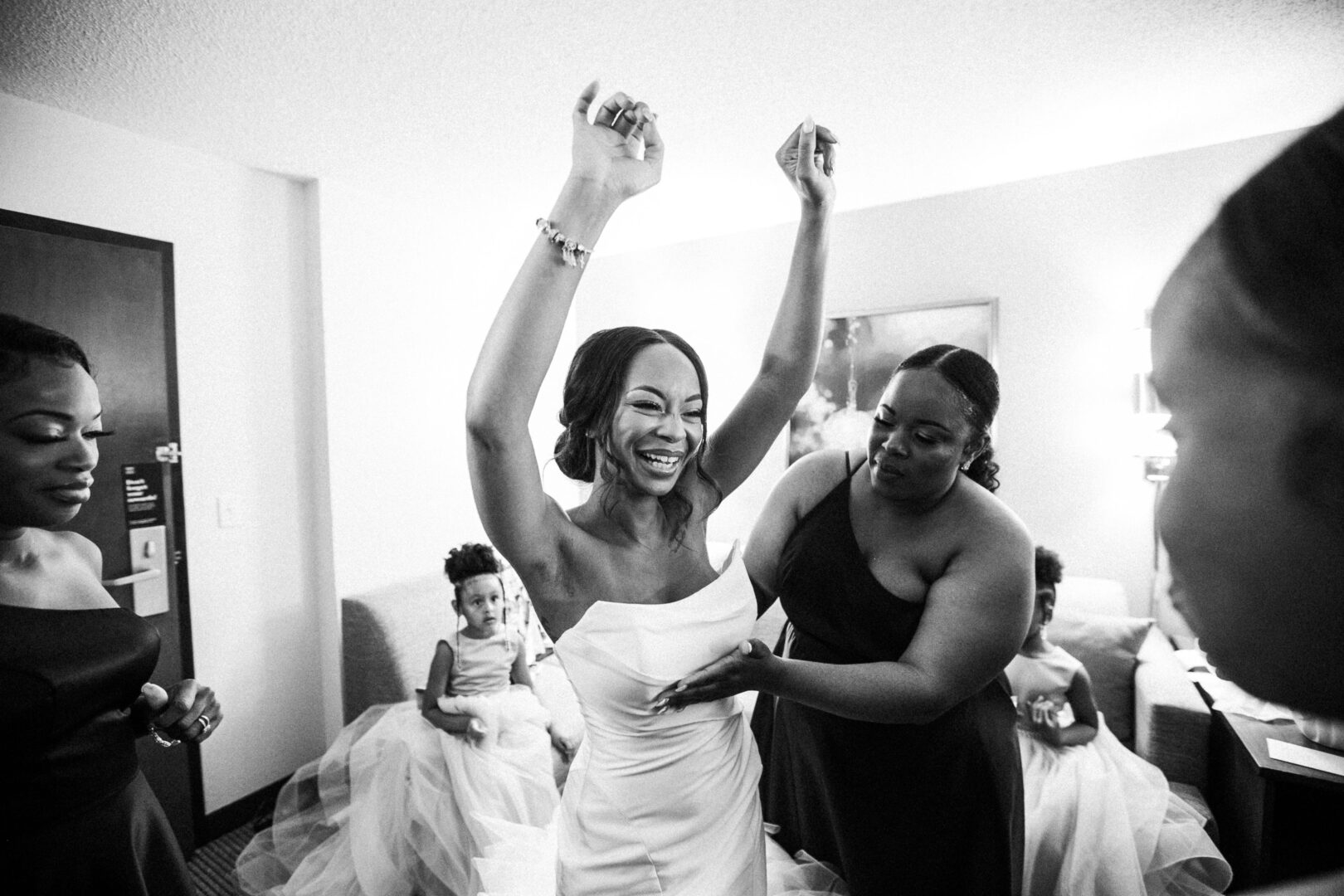 At the Mansion on Main Street Wedding, a bride joyfully celebrates with her arms raised as attendants adjust her dress, surrounded by bridesmaids in the background.