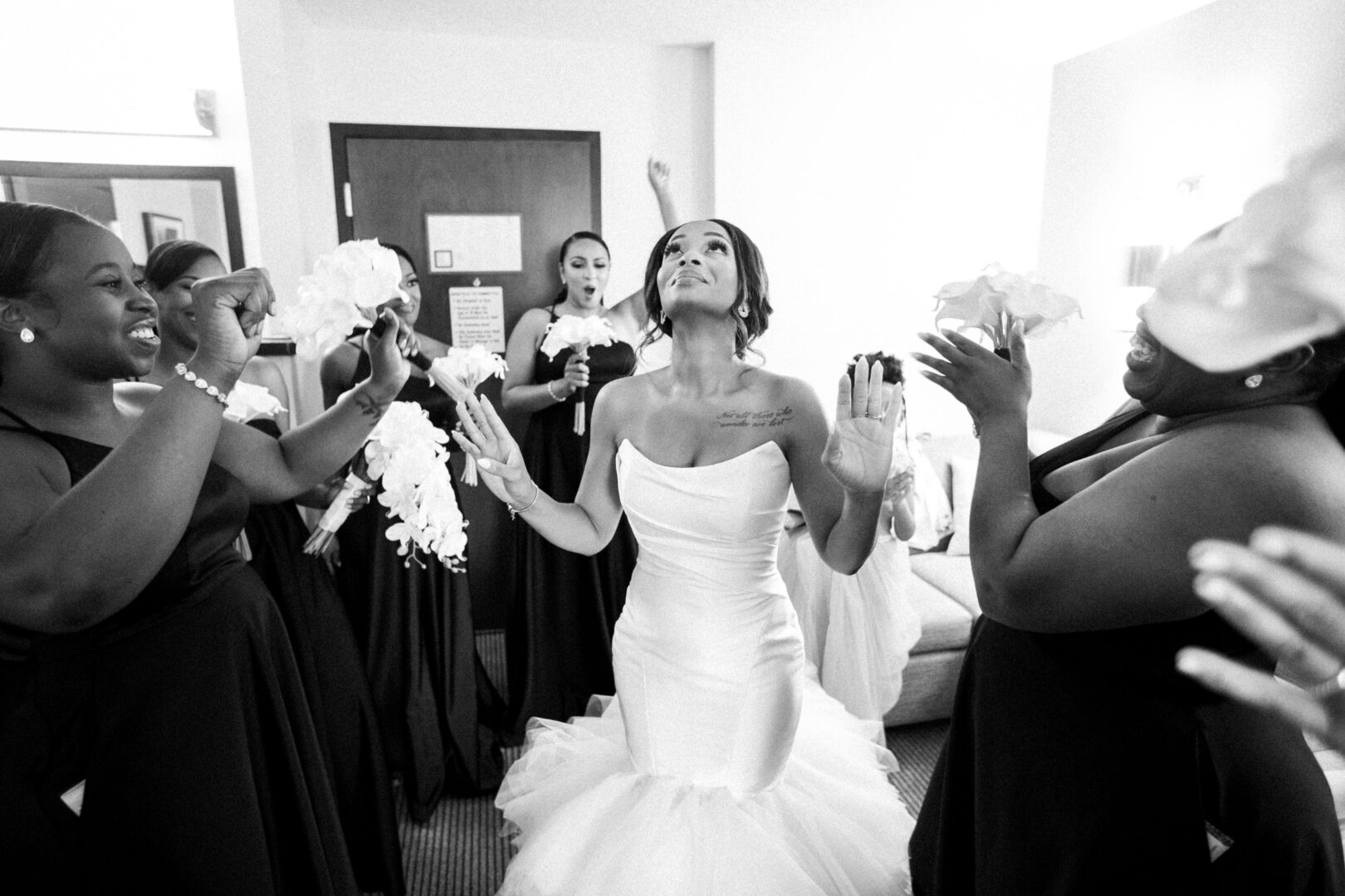 A bride in a gown stands among bridesmaids in dark dresses at the Mansion on Main Street Wedding, holding flowers and raising her hands, appearing joyous in the well-lit room.