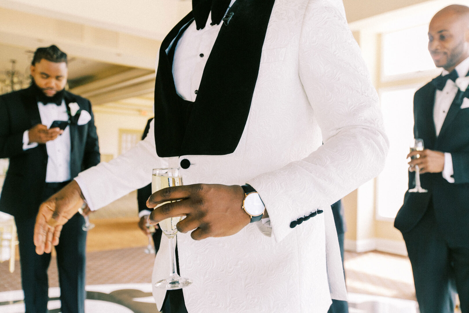 In the grandeur of the Mansion on Main Street, three men in tuxedos, one in a white jacket holding a champagne glass, stand elegantly in a bright room.
