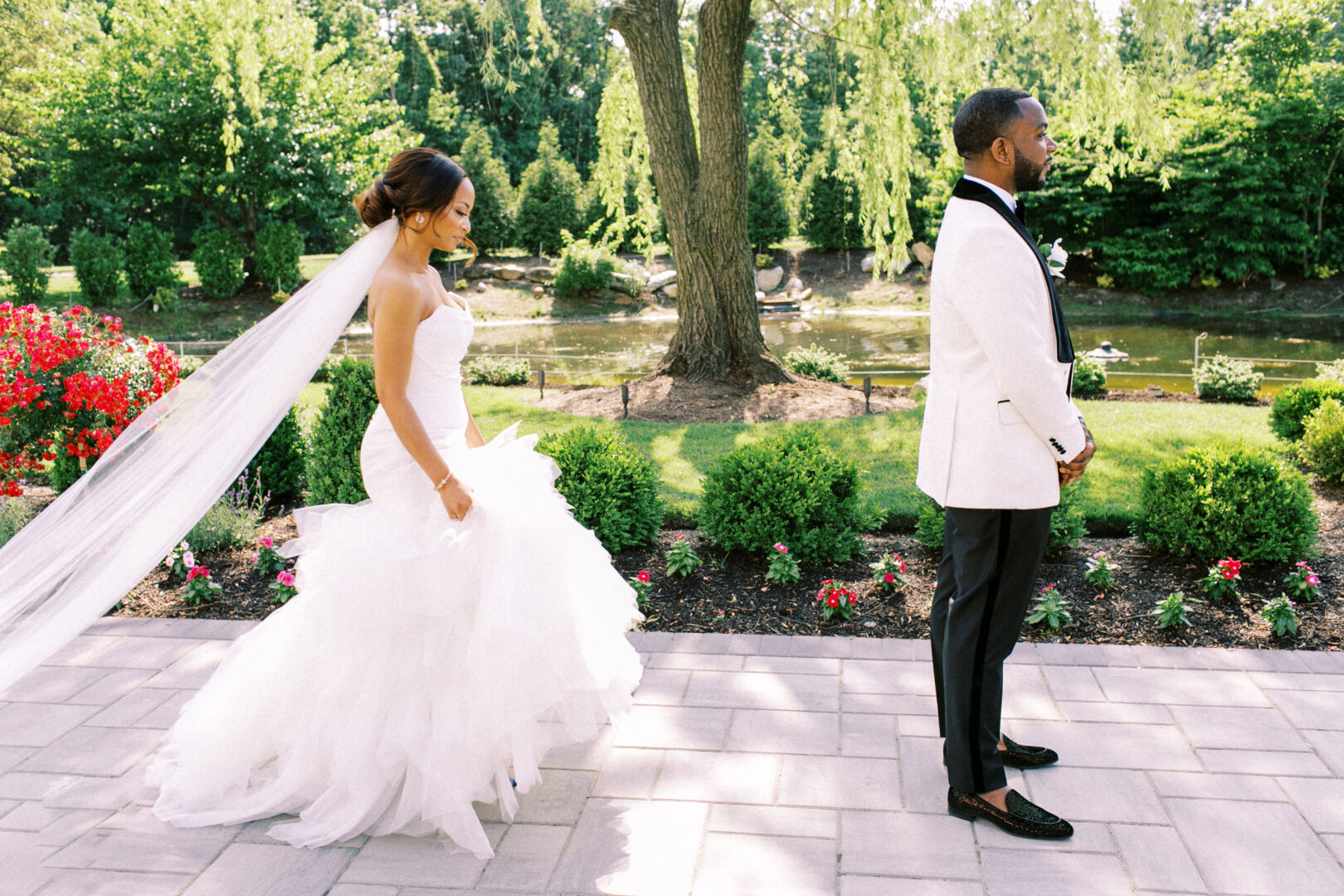 The bride in a white dress gracefully approaches a man in a white jacket and black pants on the garden path, surrounded by lush greenery and vibrant flowers at the Mansion on Main Street Wedding.