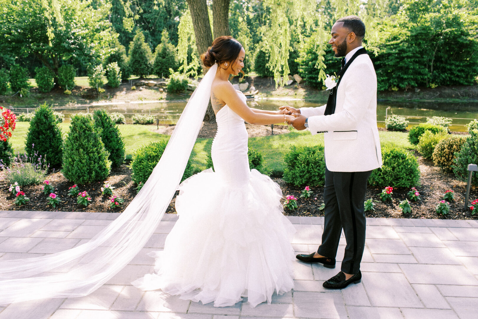 A couple in wedding attire holds hands outdoors near a pond at the elegant Mansion on Main Street, surrounded by lush greenery and vibrant flowers.