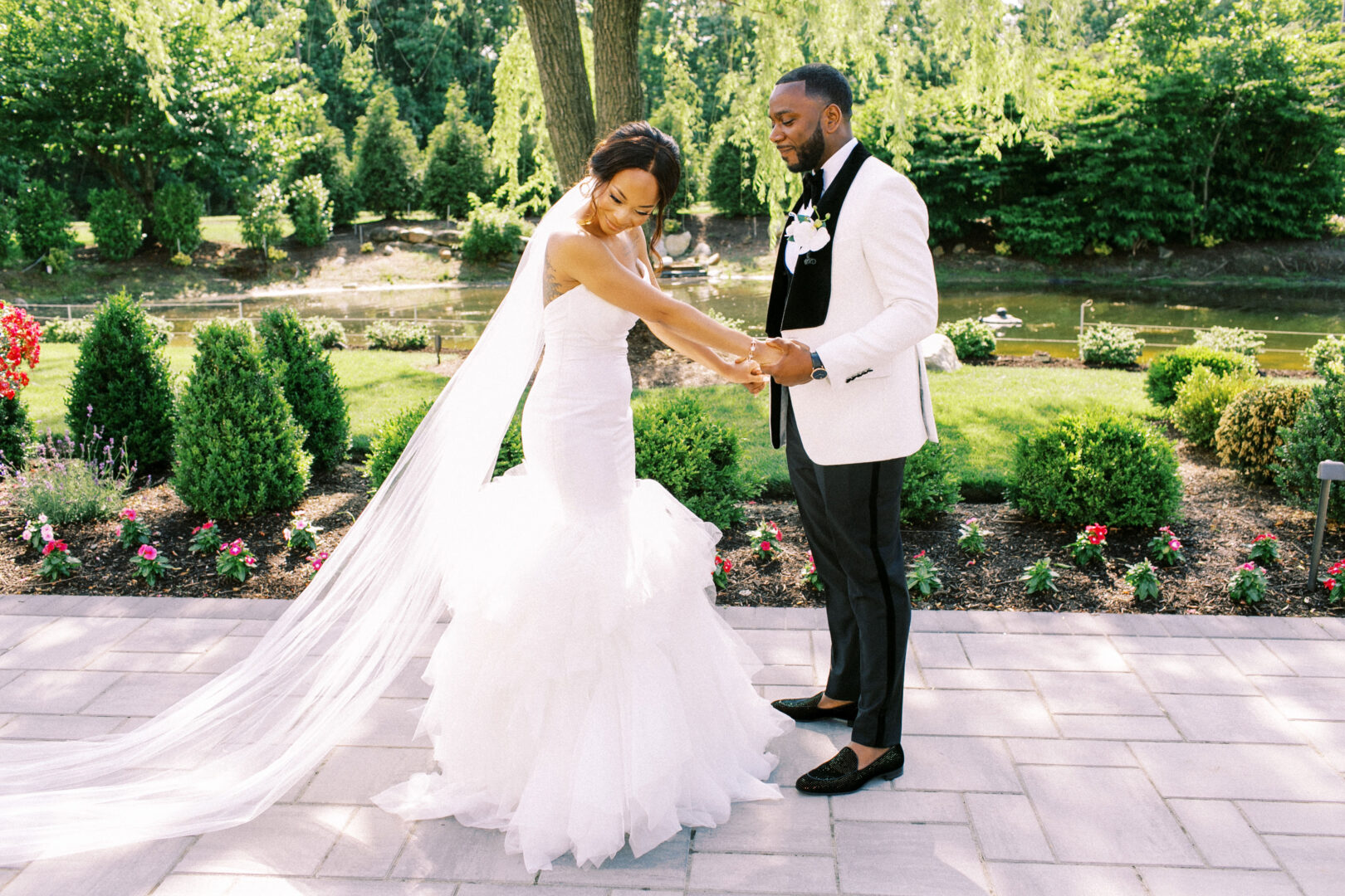 Bride and groom hold hands outdoors on a sunny day, with a garden and trees in the background at the Mansion on Main Street. The bride is in a white dress with a long veil, while the groom wears an elegant white jacket.