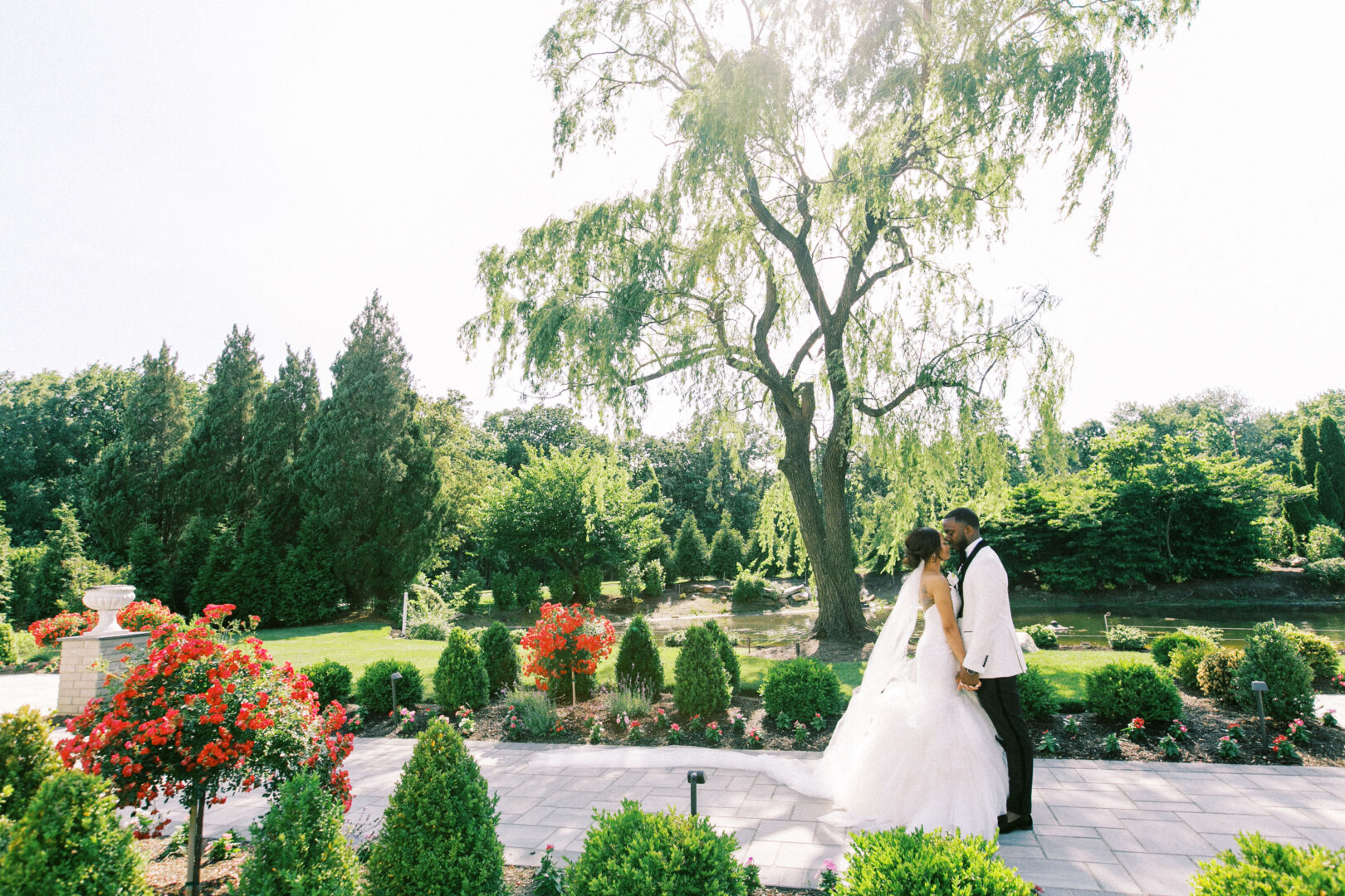 A couple in wedding attire kisses in a garden setting, surrounded by vibrant flowers and greenery, with a large tree in the background, capturing the essence of a romantic Mansion on Main Street wedding.