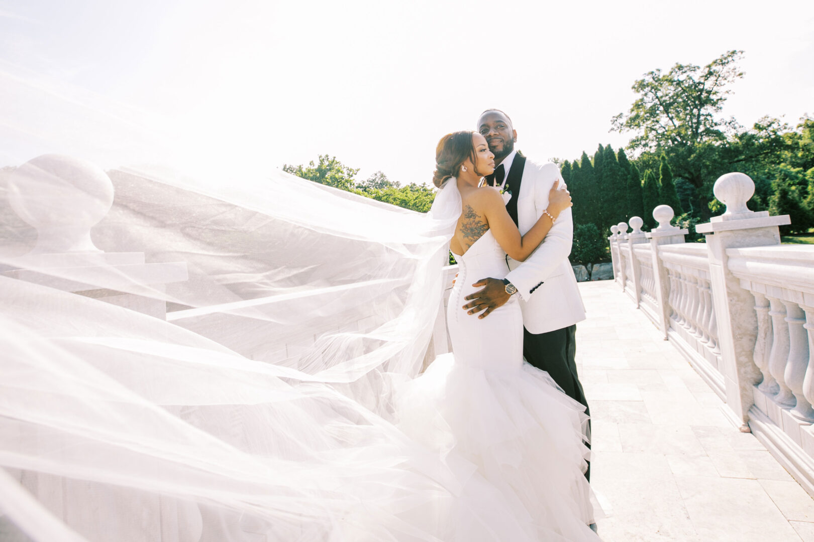 The bride and groom, in elegant wedding attire, embrace on a marble terrace at the Mansion on Main Street. The bride's veil flows gracefully in the gentle breeze, adding to the enchanting scene.