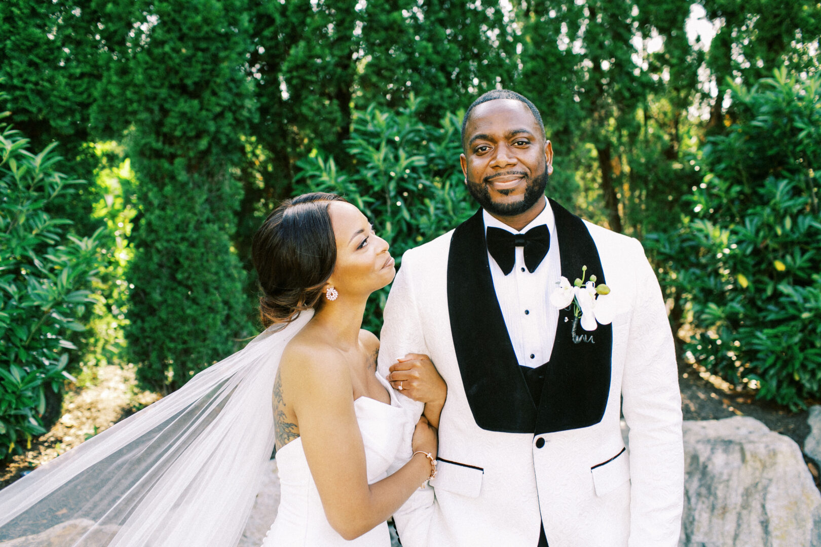 A couple stands together outdoors at the Mansion on Main Street Wedding; the woman in a white gown with a veil, and the man in a white tuxedo with black accents, surrounded by lush greenery.