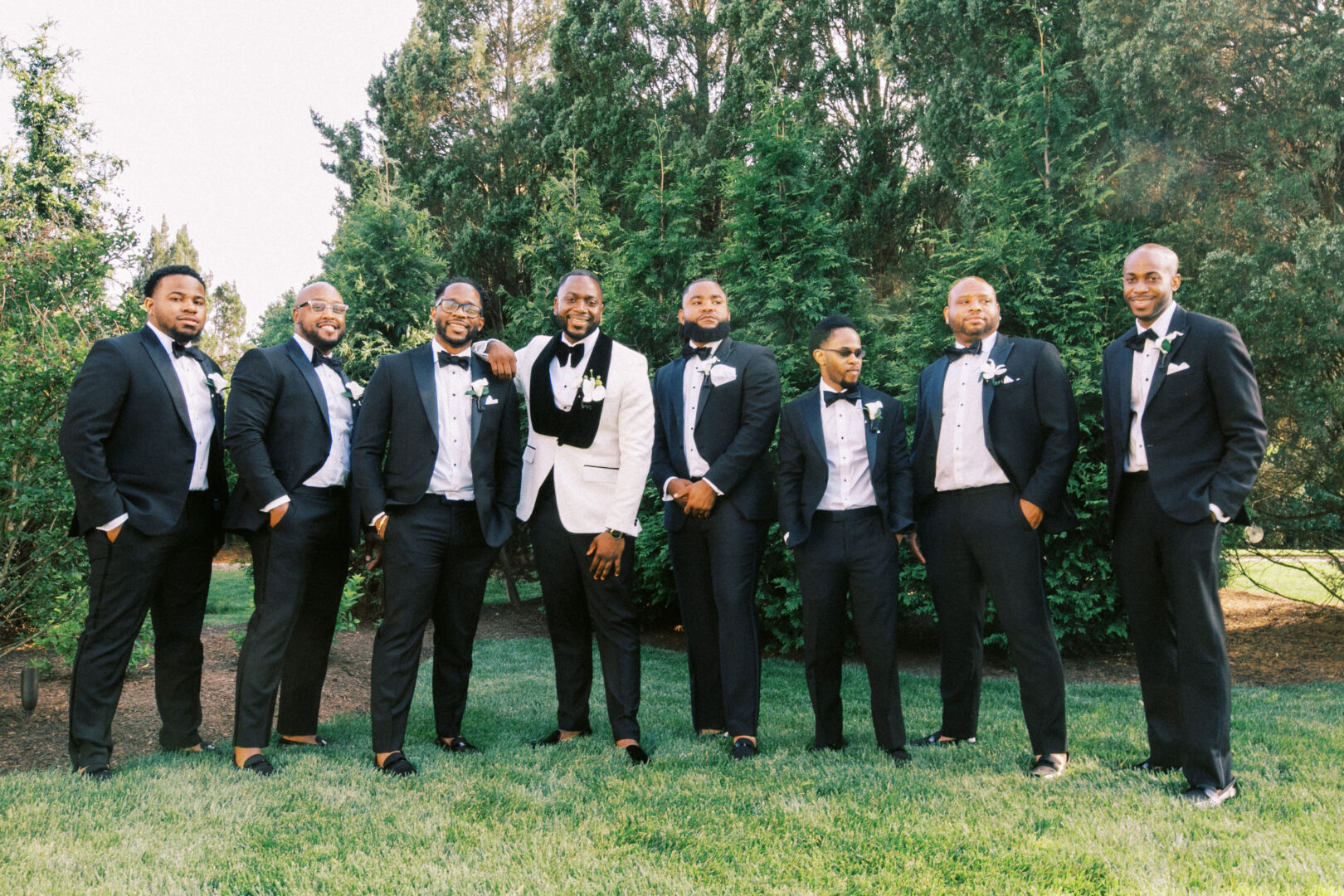 Eight men in formal black suits and bow ties, one in a white jacket, pose on the lush grass with trees behind them, capturing the elegance of a Mansion on Main Street wedding.