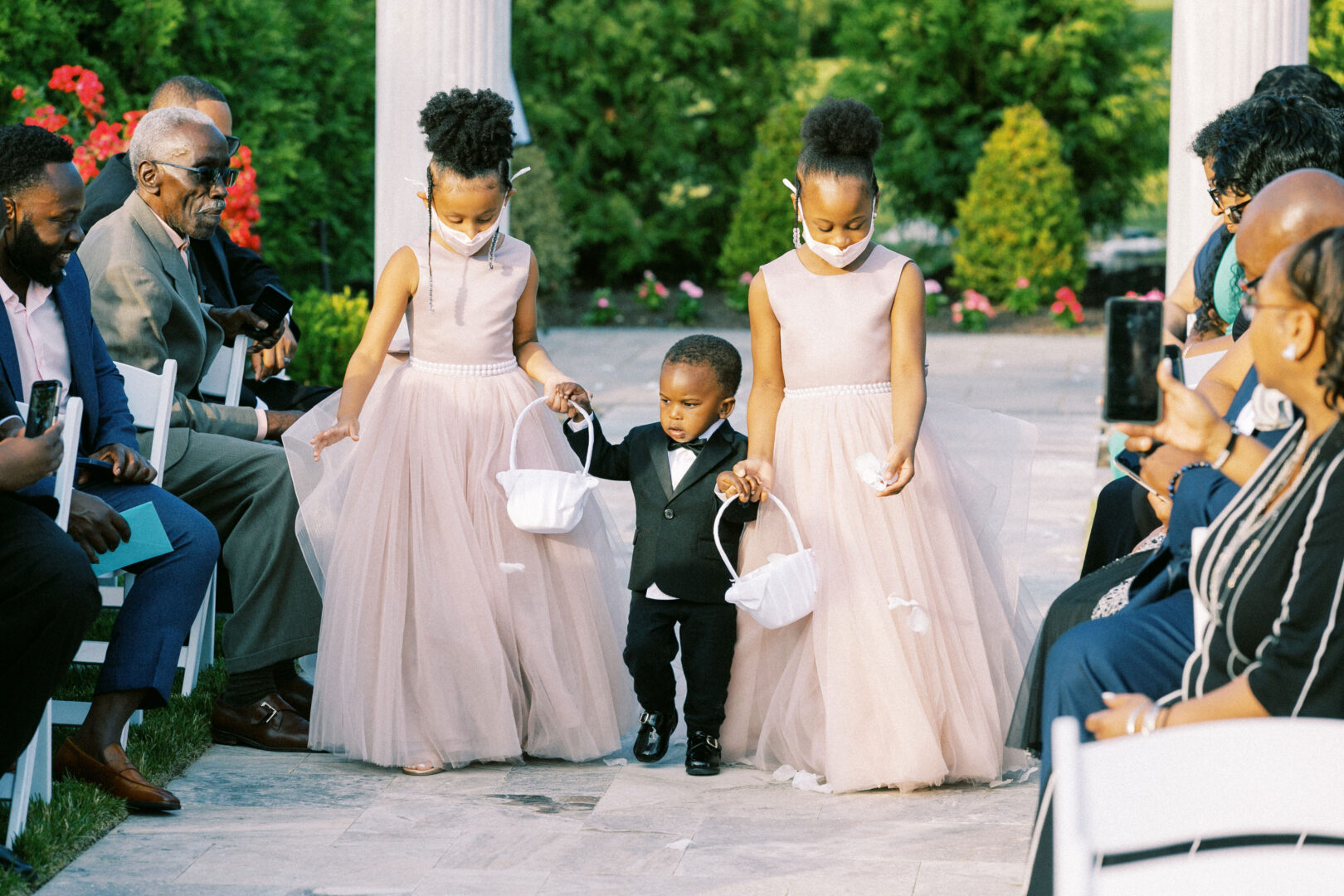 At the enchanting Mansion on Main Street wedding, three children make their way down the aisle amidst blooming gardens. Two girls in pink dresses hold flower baskets, while a boy in a suit walks between them. Guests seated nearby capture these precious moments on camera.
