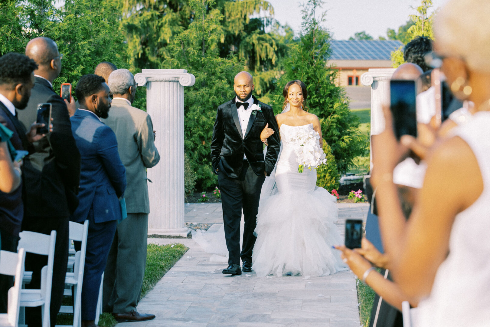 A bride in a white dress and groom in a black tuxedo walk down an outdoor aisle at the Mansion on Main Street Wedding, surrounded by seated and standing guests taking photos.