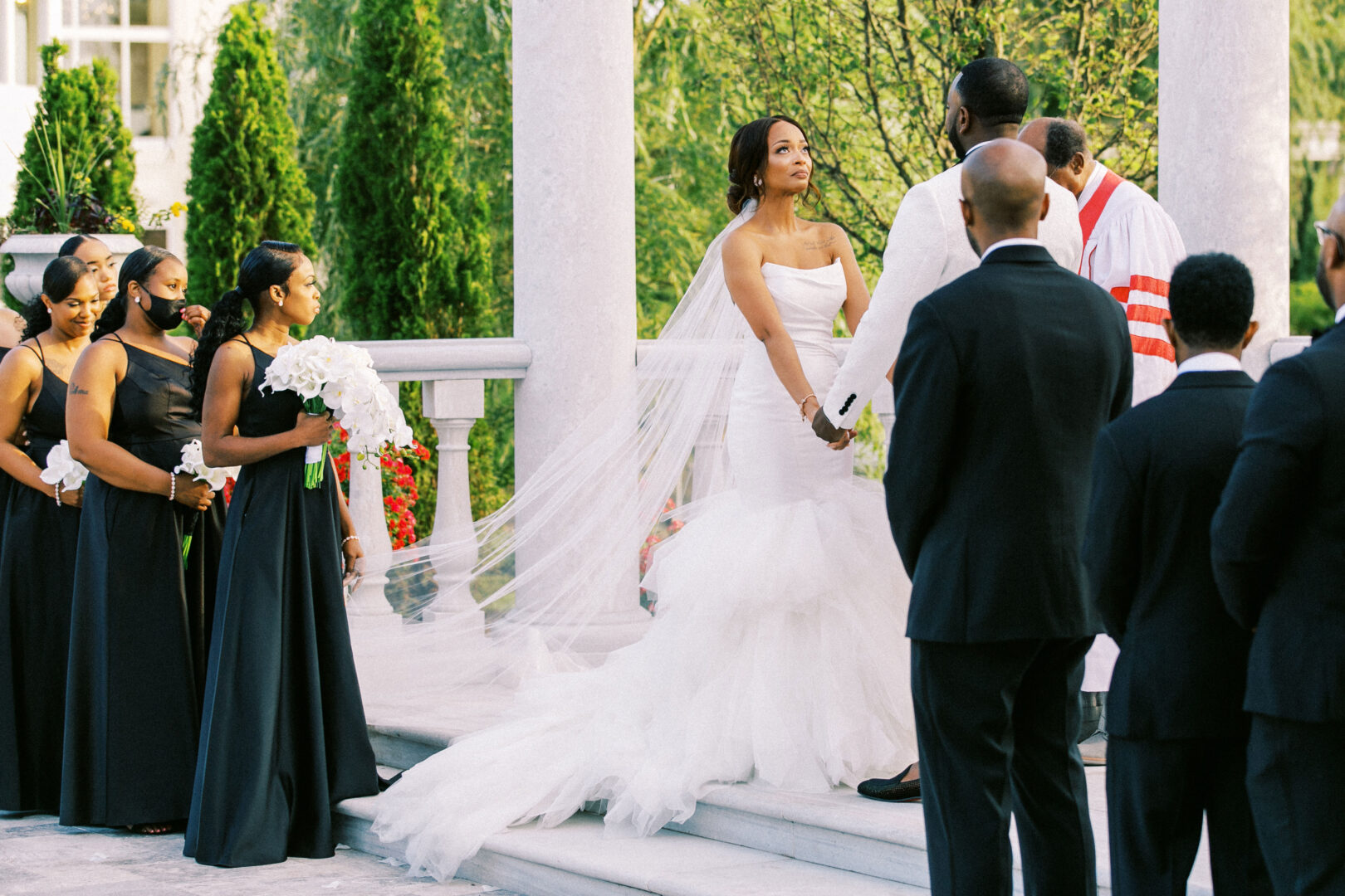 At the enchanting Mansion on Main Street, a bride and groom stand holding hands during their outdoor wedding ceremony, surrounded by bridesmaids and groomsmen.