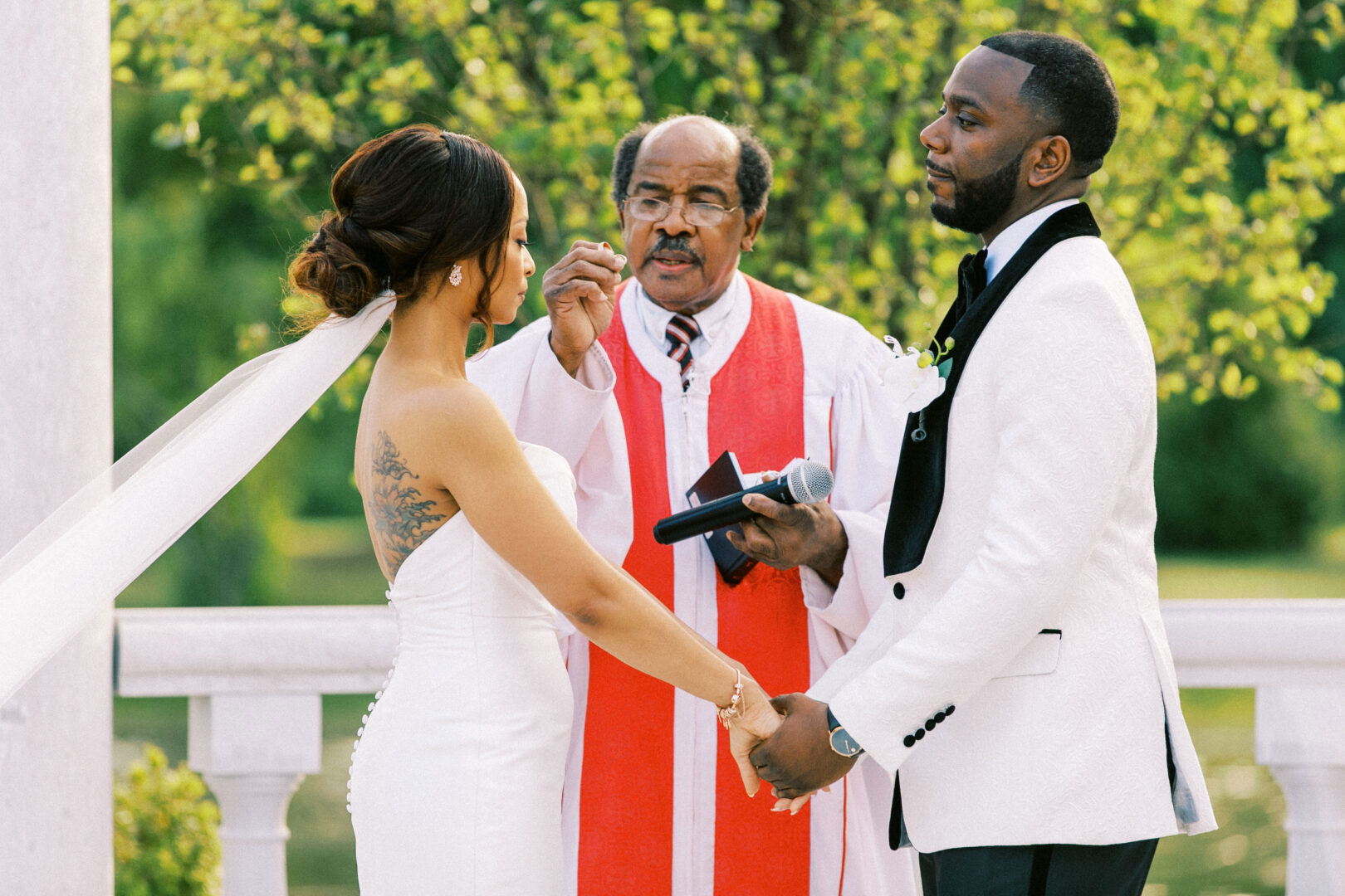 A couple holds hands during a Mansion on Main Street wedding ceremony, with an officiant in a red robe speaking behind them.