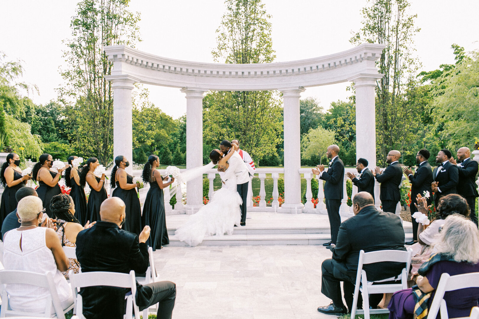 At the elegant Mansion on Main Street wedding, a couple kisses under the open sky as guests seated and standing around them offer hearty applause.
