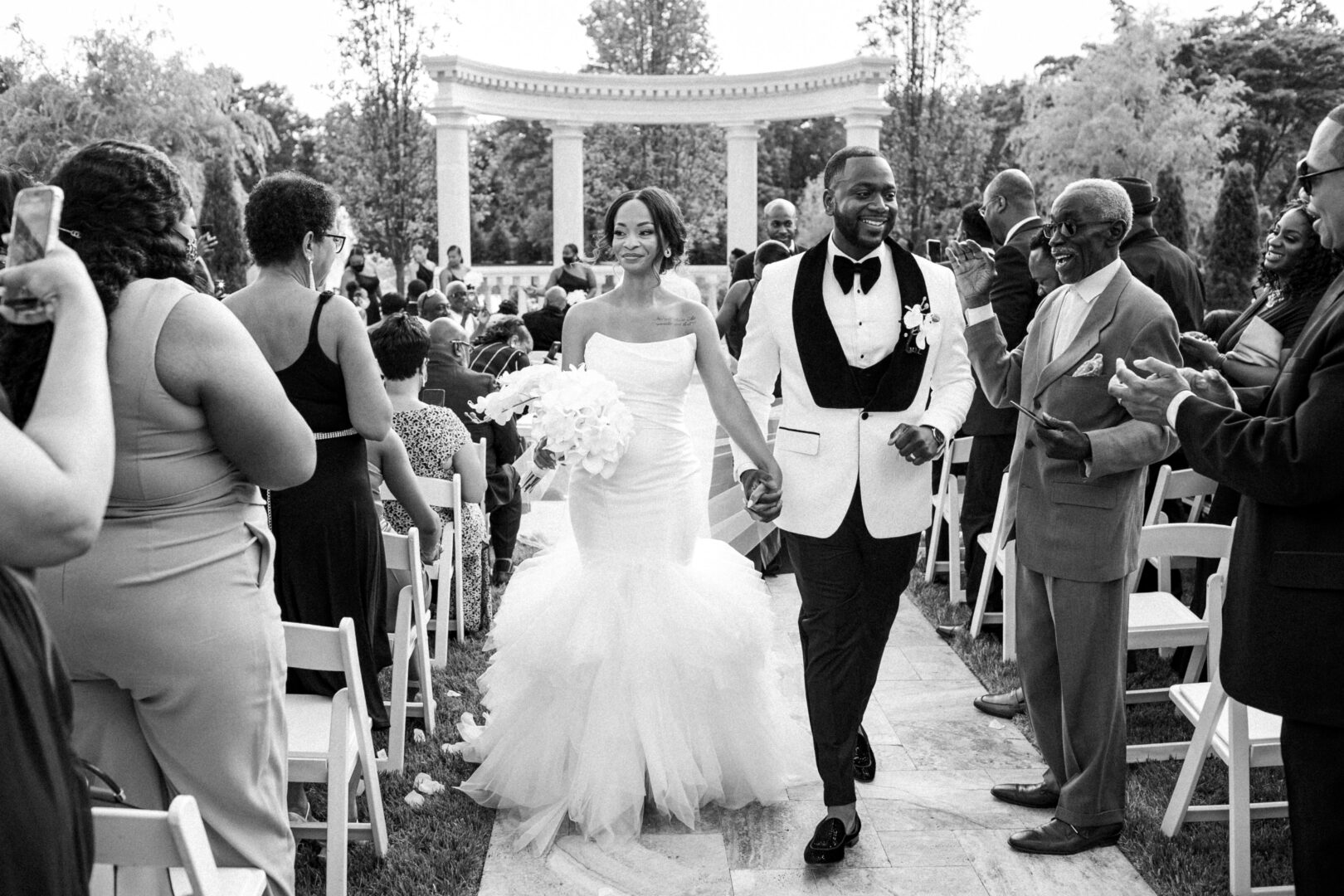 At the Mansion on Main Street, a bride and groom joyfully walk down the aisle, hand in hand, as guests applaud. The elegant outdoor setting features a stunning archway in the background.