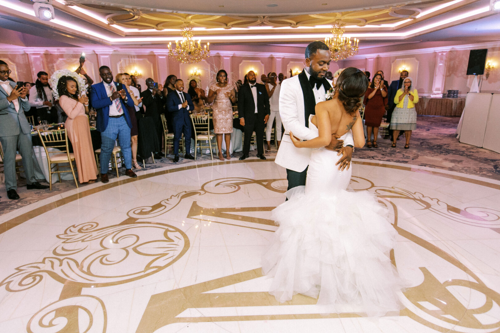 A couple shares their first dance at a Mansion on Main Street wedding reception, surrounded by guests dressed in formal attire, in an elegantly decorated ballroom.