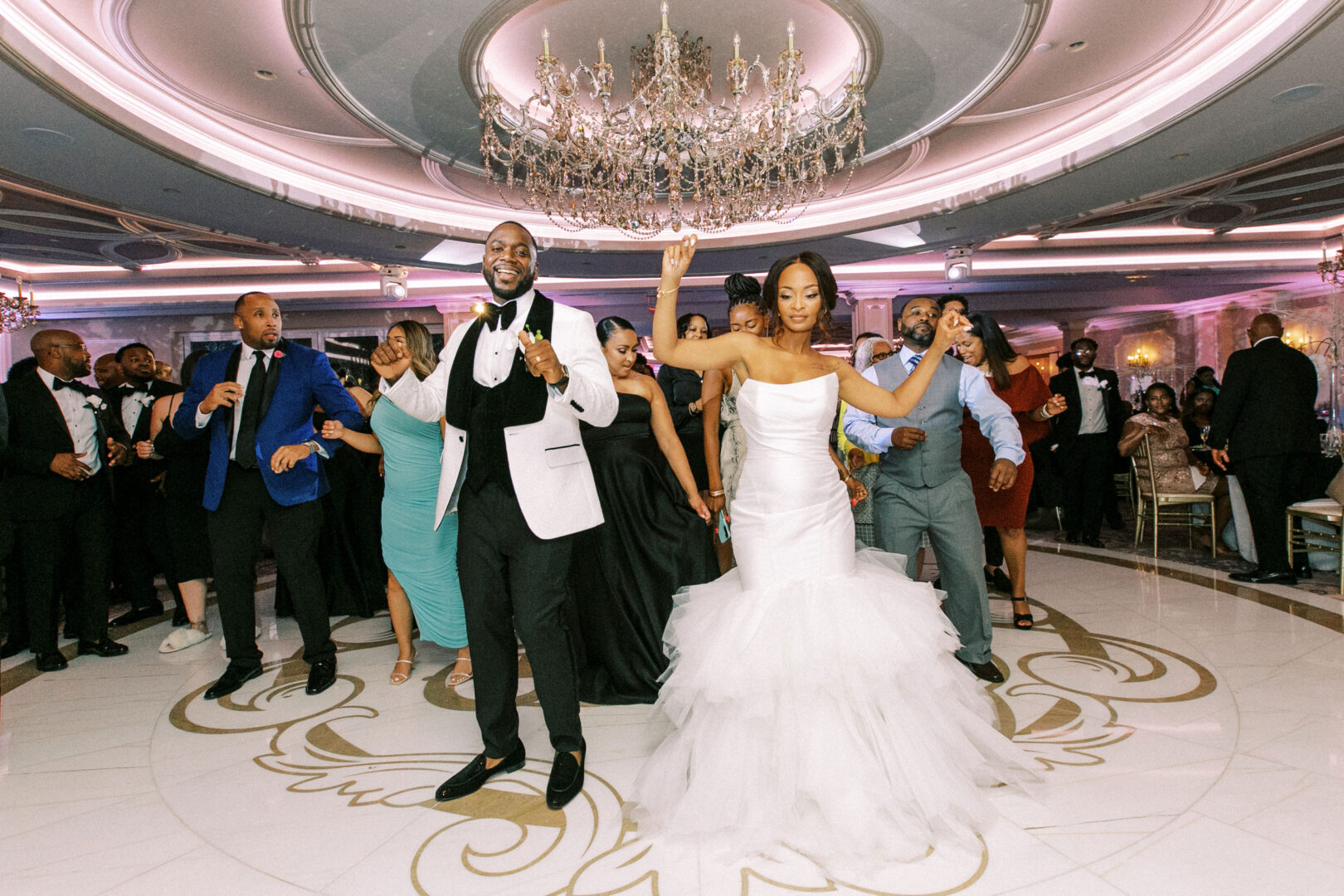 A bride and groom joyfully dance with their guests on the elegant ballroom floor of the Mansion on Main Street, illuminated by a large chandelier.