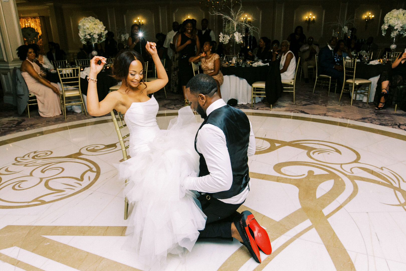 At a charming Mansion on Main Street Wedding, a groom kneels gracefully to remove a garter from his seated bride's leg, while delighted guests watch this timeless tradition unfold in the elegant setting.