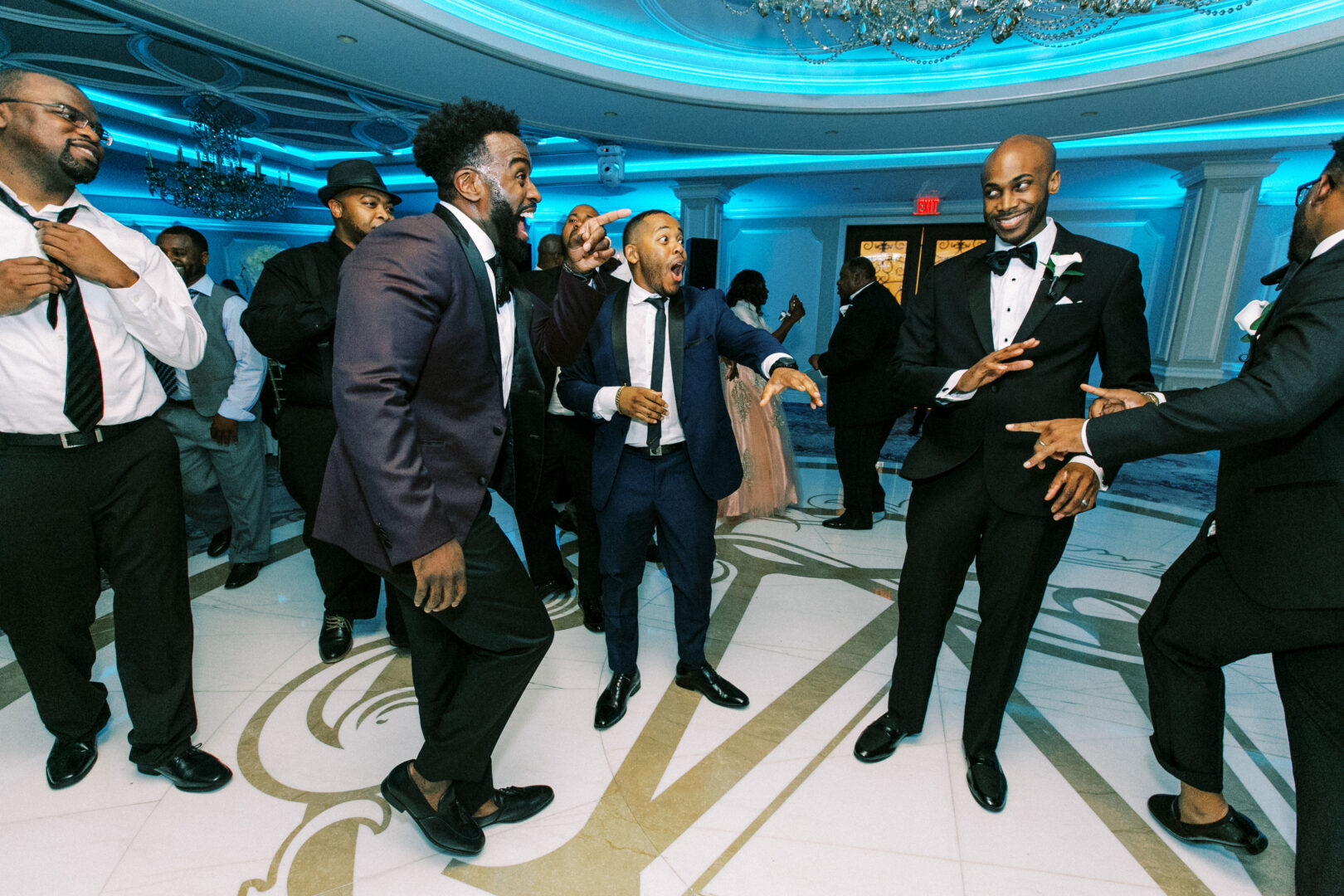 A group of men in formal attire are laughing and dancing in the brightly lit ballroom of the Mansion on Main Street Wedding venue, with a chandelier overhead and blue ambient lighting setting the perfect celebratory mood.