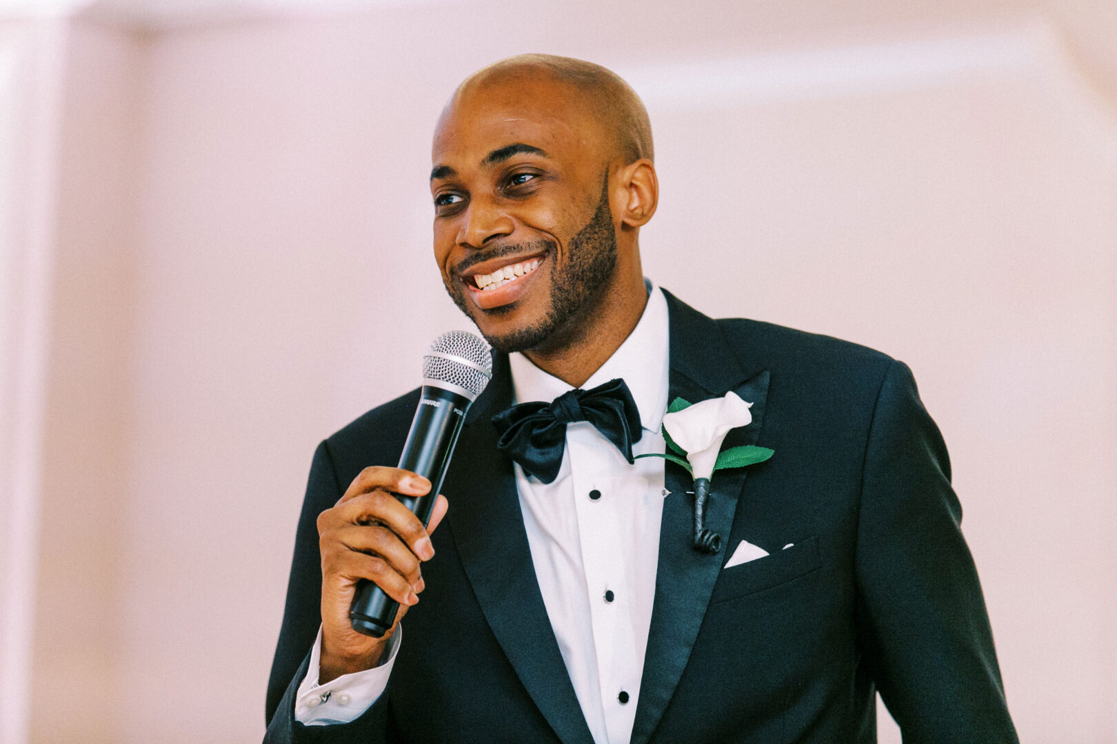 A man in a suit holds a microphone and smiles while speaking at the Mansion on Main Street Wedding, adding charm and elegance to the enchanting celebration.
