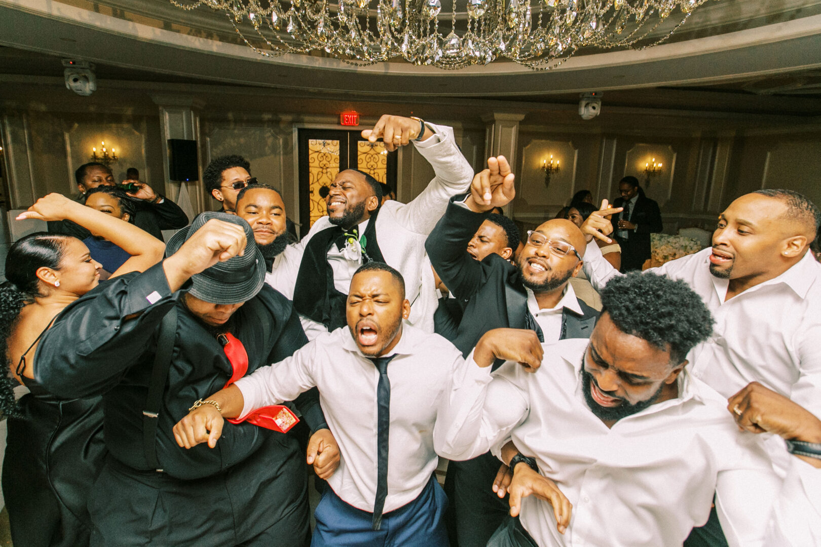 A group of people energetically dancing and cheering beneath chandeliers in the well-lit ballroom of the Mansion on Main Street.