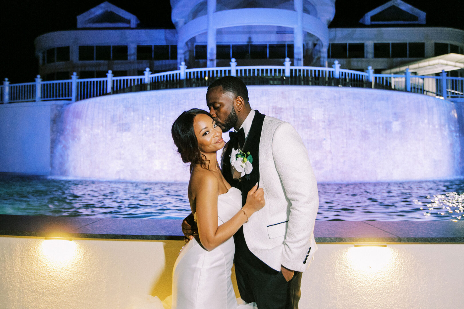 A bride and groom stand in front of a lit-up fountain at night during their Mansion on Main Street Wedding. The groom kisses the bride's forehead while she smiles at the camera.