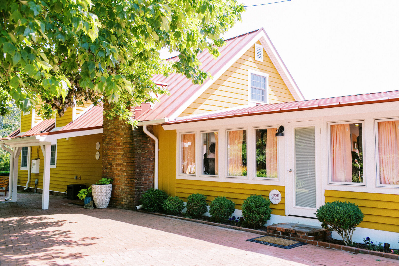 A yellow house with a red roof, large windows, and a brick chimney exudes charm reminiscent of a Willow Creek Winery wedding. Surrounded by green bushes and a tree, it sits gracefully beside a brick-paved driveway.