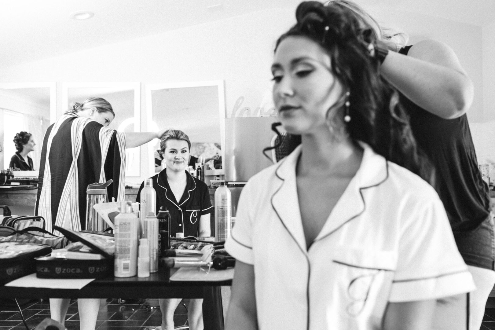 Two women are getting their hair styled in a salon, wearing matching pajamas, as they prepare for the Willow Creek Winery wedding. One is seated in front of a mirror while the other sits in the foreground, both excited for the day ahead.