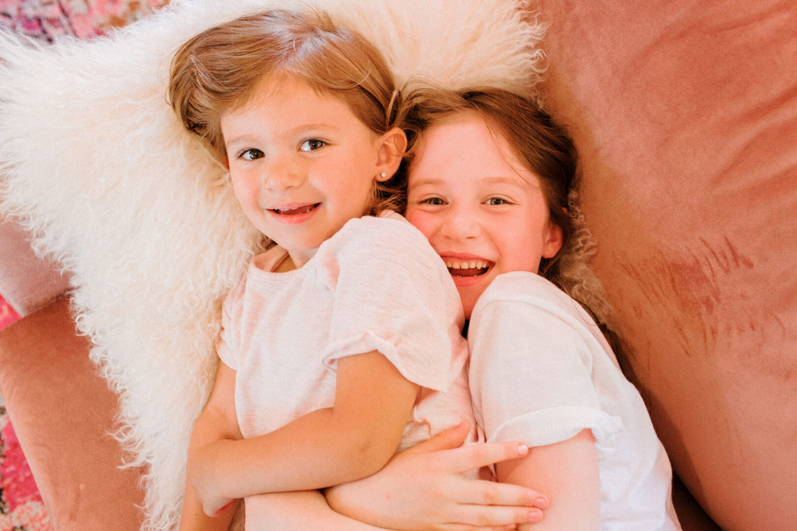 Two young children lying on a pink sofa, smiling and hugging, with a fluffy white pillow behind them, reminiscent of the warmth and joy found at Willow Creek Winery weddings.