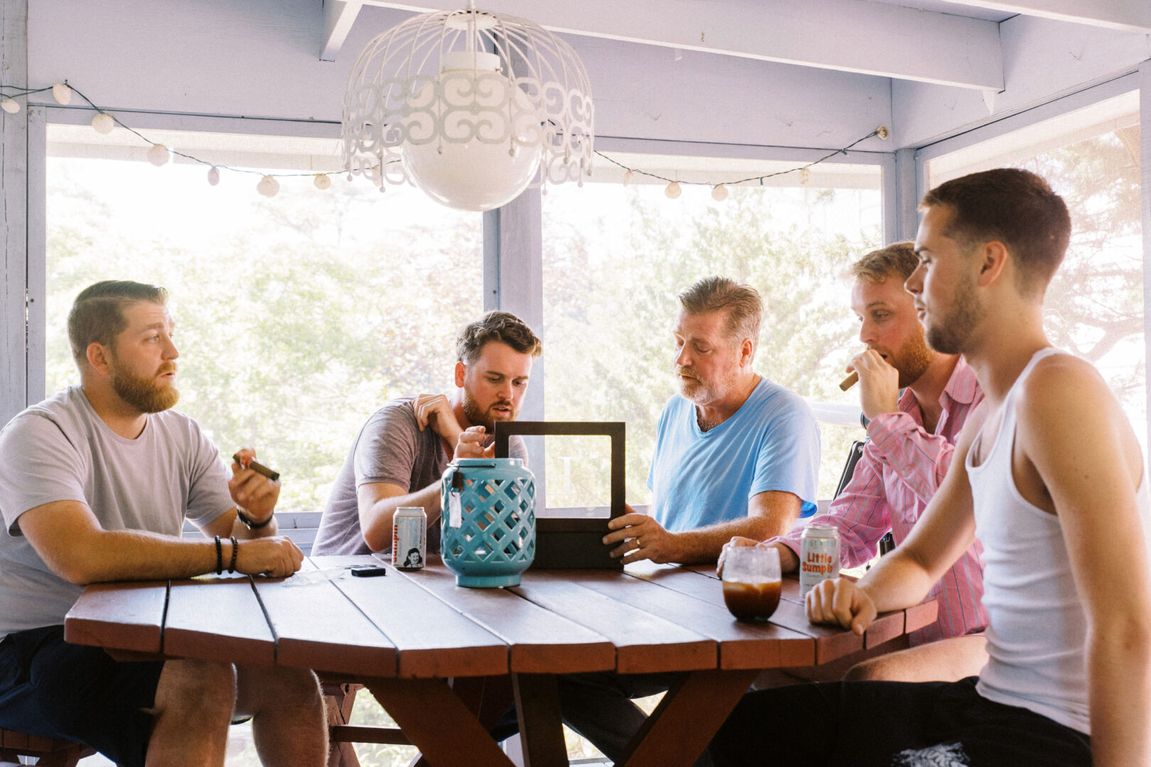 Five men sit around a wooden table on a porch at Willow Creek Winery, engaging in lively conversation. Drinks and a blue decorative lantern are on the table, setting the perfect atmosphere for a memorable wedding moment.