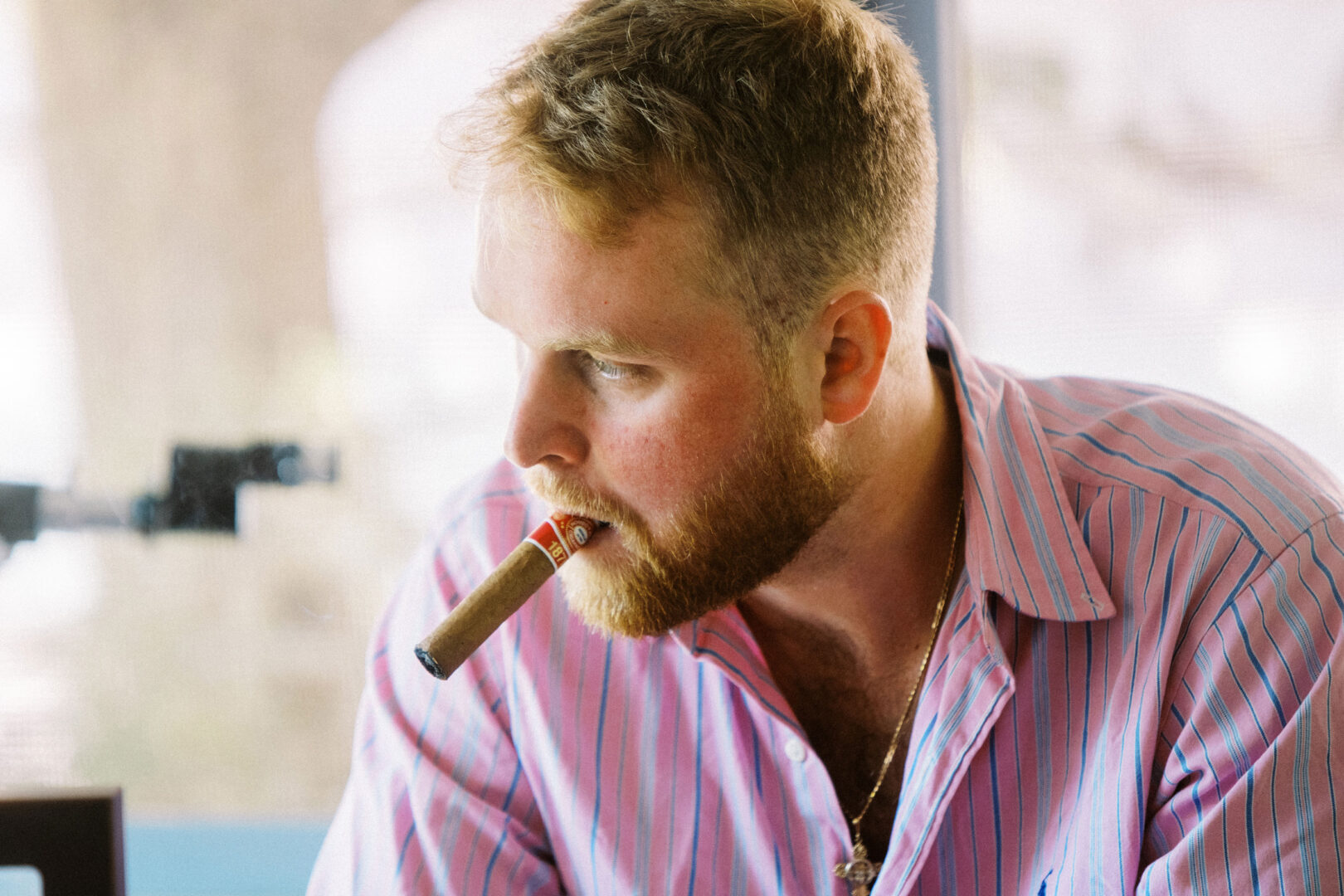 A man in a pink striped shirt, possibly from a recent Willow Creek Winery wedding, holds a cigar between his lips as he gazes to the side.