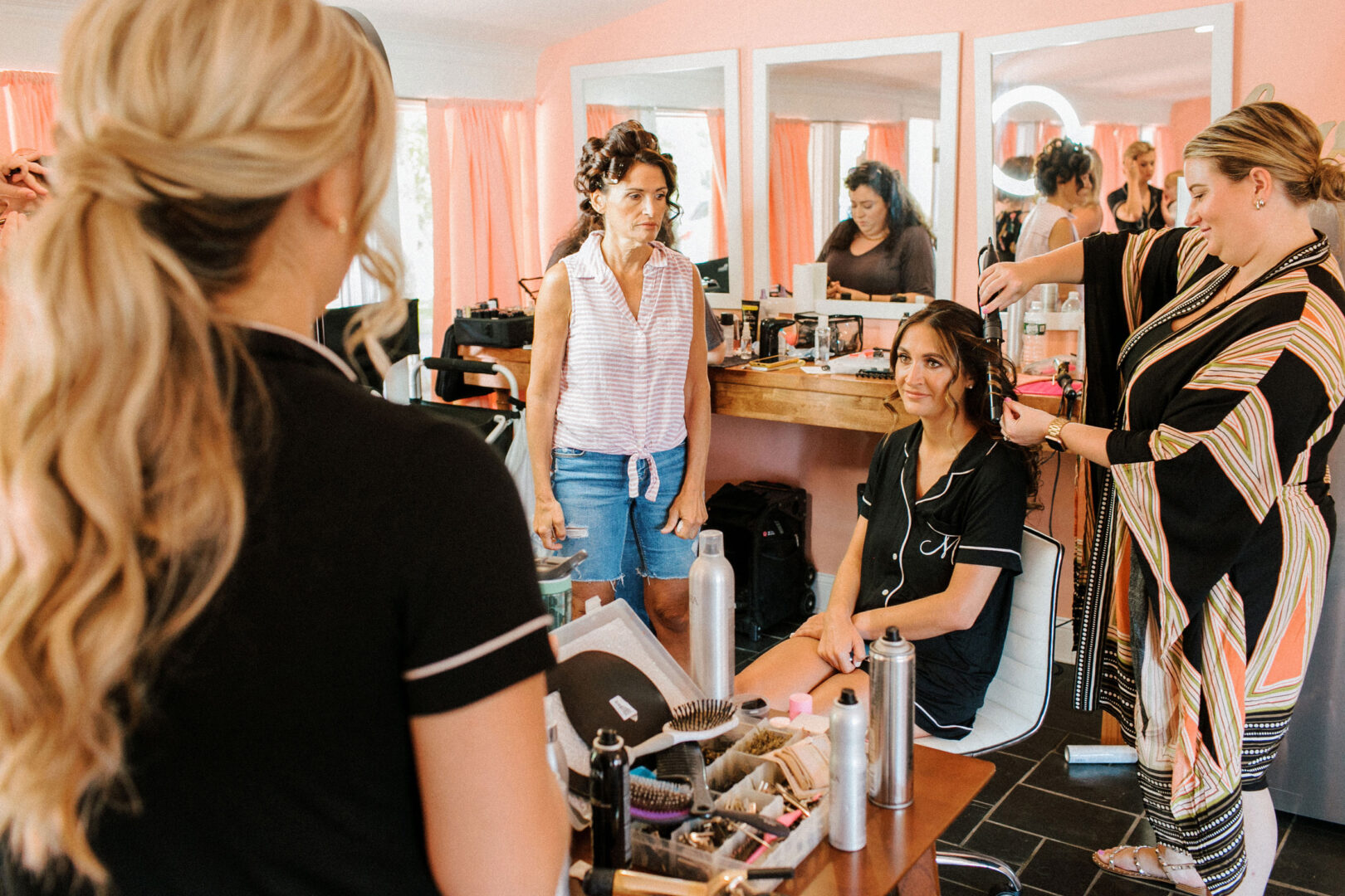 Several women are in a room with pink walls at Willow Creek Winery. One woman is seated, getting her hair styled, while others stand nearby with hair and makeup supplies on a table, preparing for the wedding.