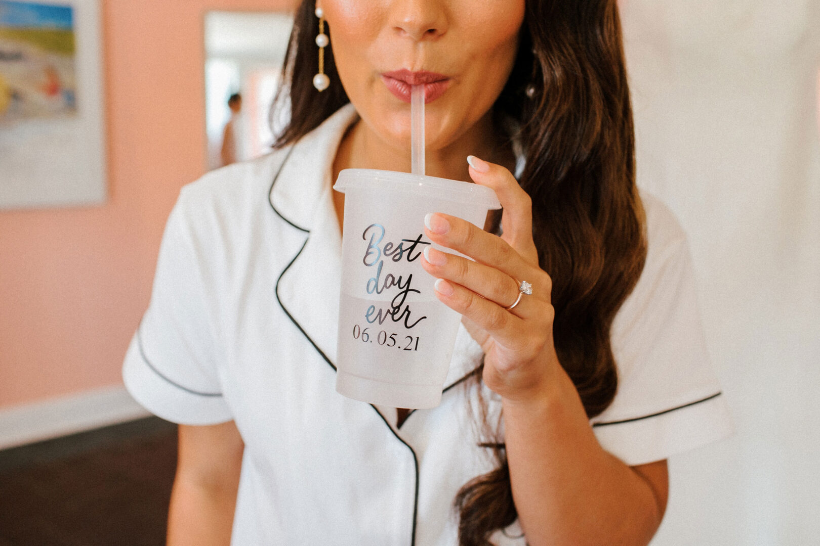 A woman in a white shirt sips from a clear cup labeled "Best day ever 06.05.21," wearing pearl earrings and a visible engagement ring, reminiscent of the joyous moments at Willow Creek Winery Wedding.