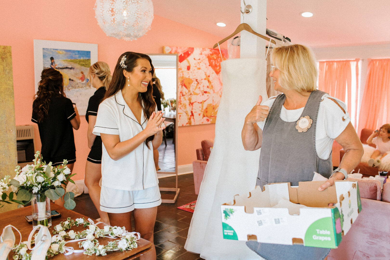 A woman in pajamas chats with another holding a fruit box, framed by the elegance of a Willow Creek Winery wedding. Nearby, a wedding dress hangs gracefully as a bridesmaid adjusts flowers in the background.