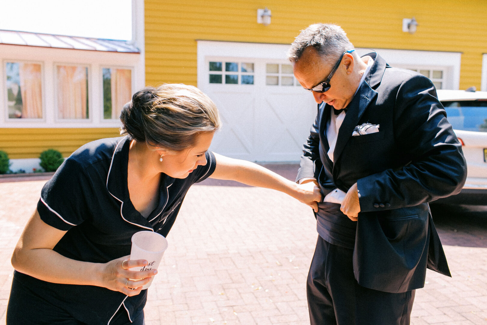 A woman helps adjust a man's suit jacket outdoors at the Willow Creek Winery wedding. He wears sunglasses, and she holds a cup. They stand in front of a yellow building with white doors, capturing a moment of elegance and charm amidst the vineyard's scenic backdrop.