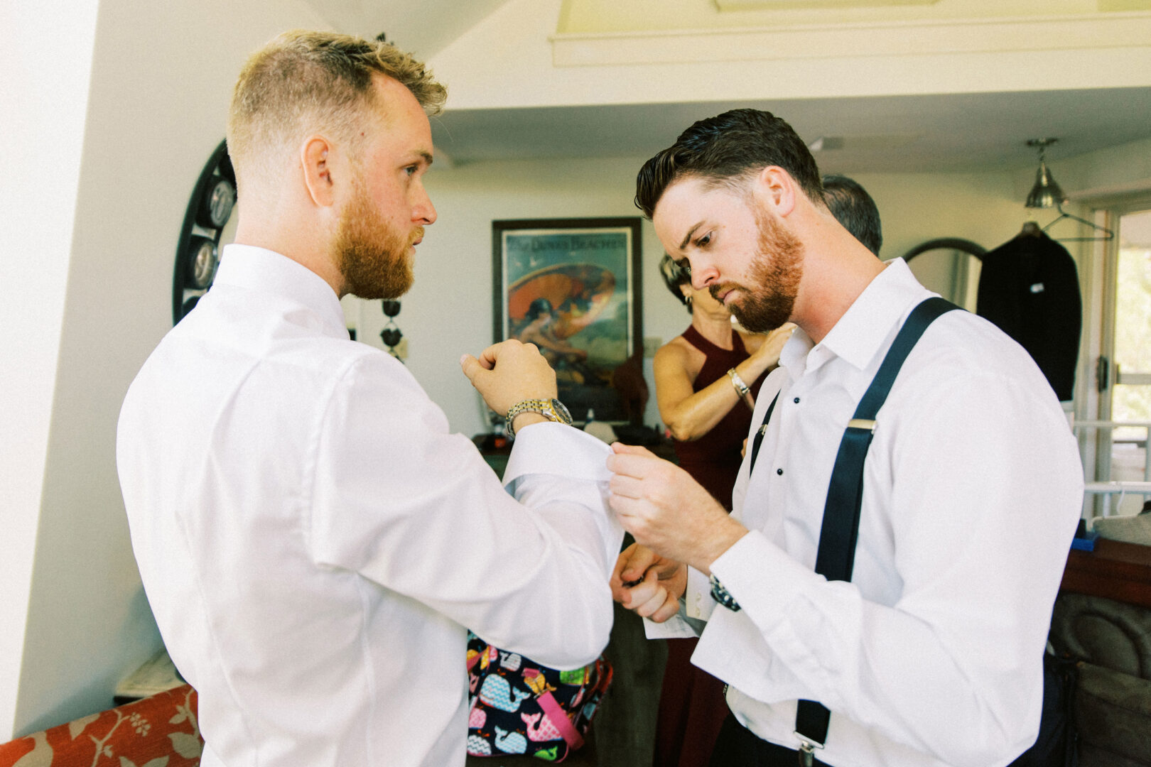 Two men in dress shirts adjust their cufflinks in a room adorned with clothing and a poster, preparing for the Willow Creek Winery wedding. A woman stands in the background, adding to the scene's anticipation.