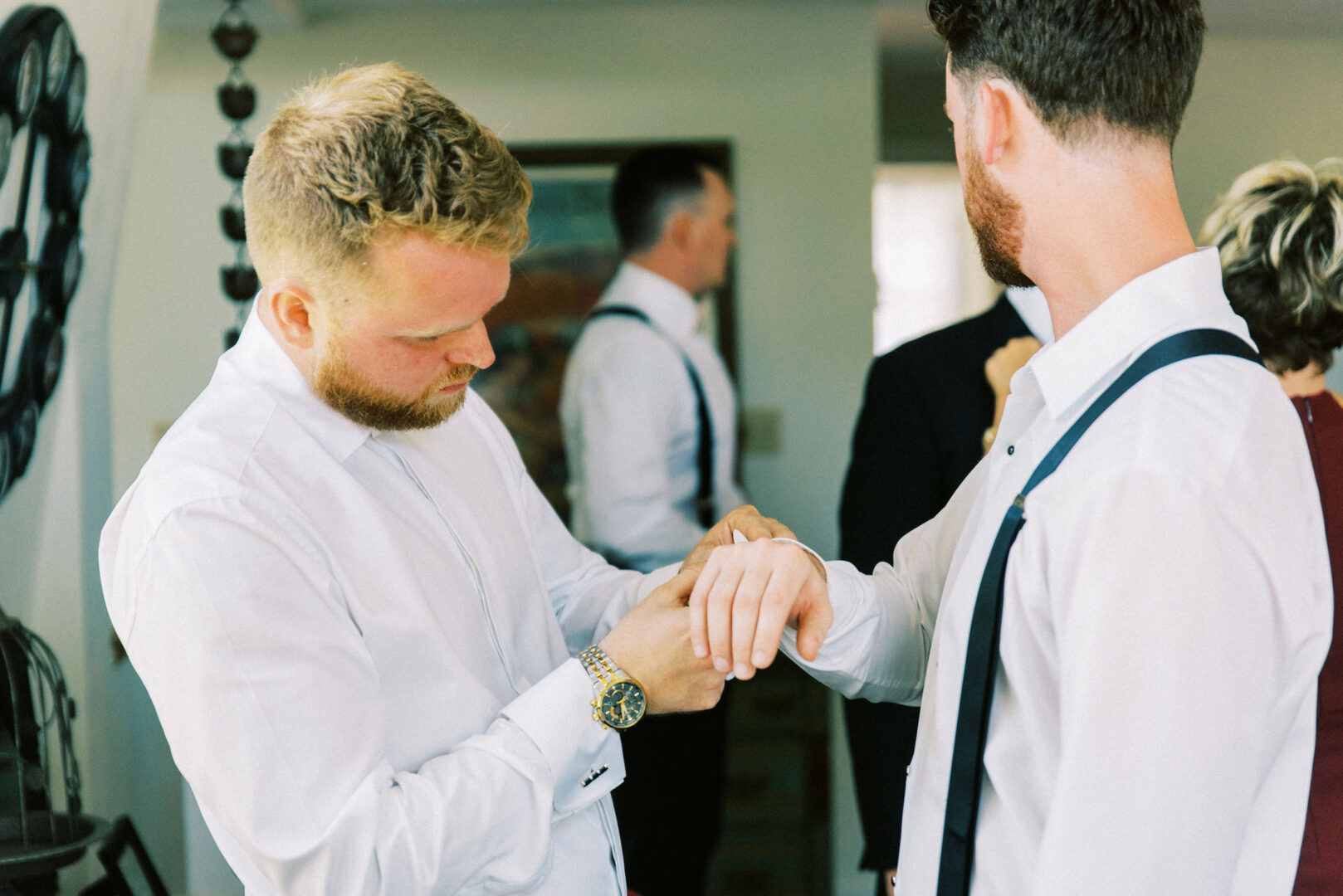 Two men in white shirts adjust each other's cuffs in a room bustling with guests at the Willow Creek Winery wedding, a beautifully decorated backdrop highlighting the joyous occasion.