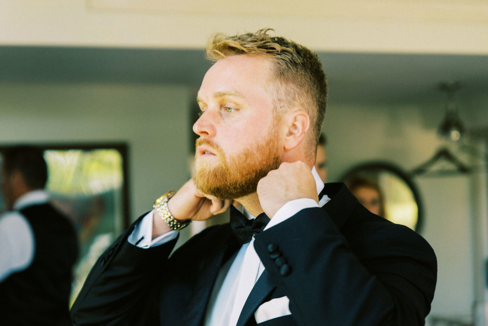 In a well-lit room, a man adjusts his black bow tie, dressed impeccably in a black suit and white shirt, preparing for the elegance of a Willow Creek Winery wedding.