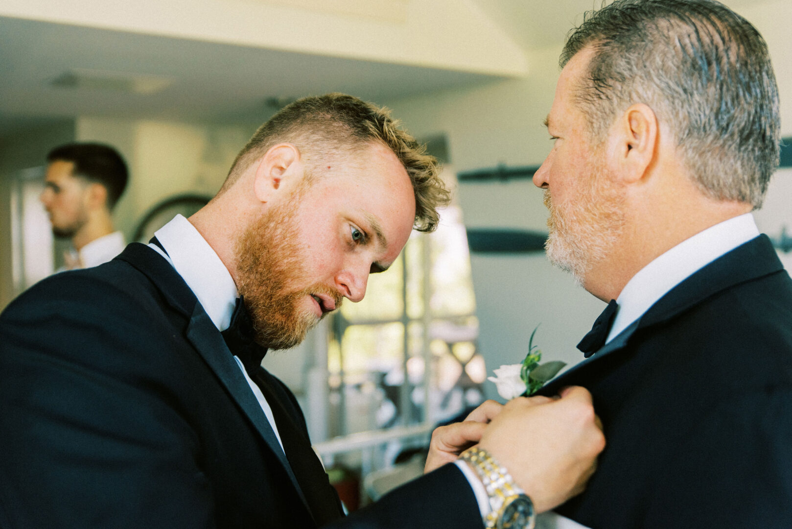 A bearded man in a tuxedo adjusts a boutonniere on another man's suit, the serene charm of Willow Creek Winery Wedding evident as someone stands in the background.