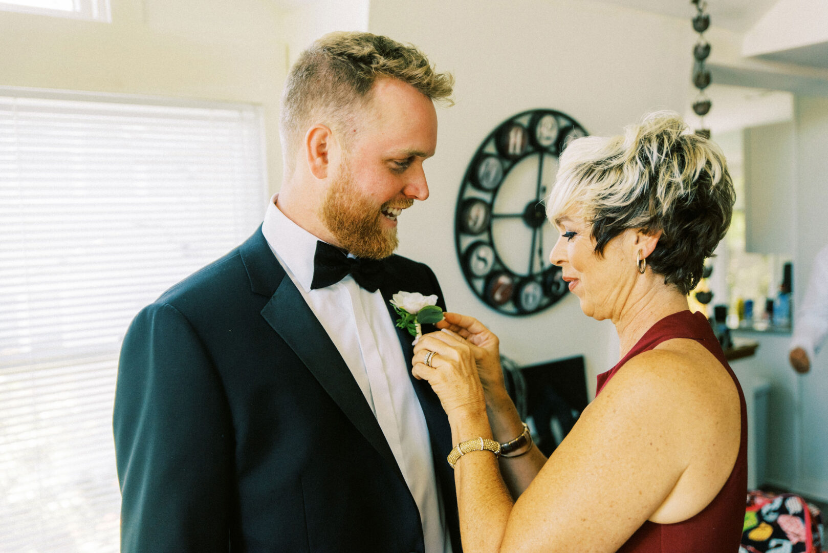 A woman pins a boutonniere on a man's suit jacket during their Willow Creek Winery wedding. They are indoors, and the man is wearing a black tuxedo with a bow tie. A round wall clock is visible in the background, capturing the elegance of the moment.