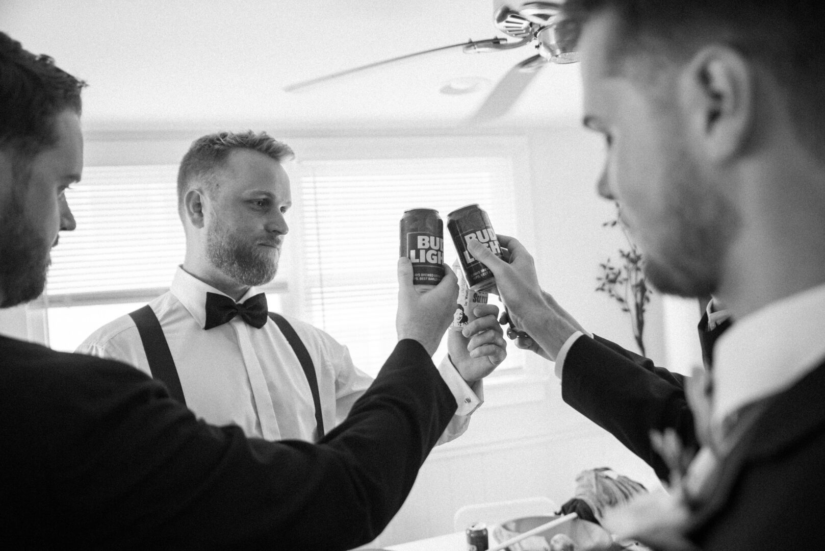 A group of men in formal attire is raising a toast with canned drinks, celebrating in the bright room of a Willow Creek Winery wedding.