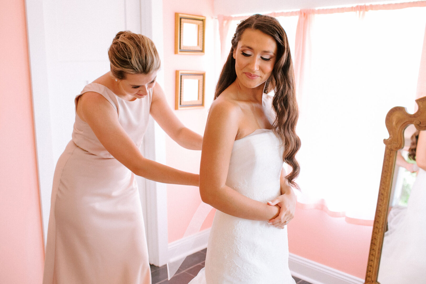 In a brightly lit room at Willow Creek Winery, a woman in a pink dress helps another woman in a bridal gown adjust her dress.