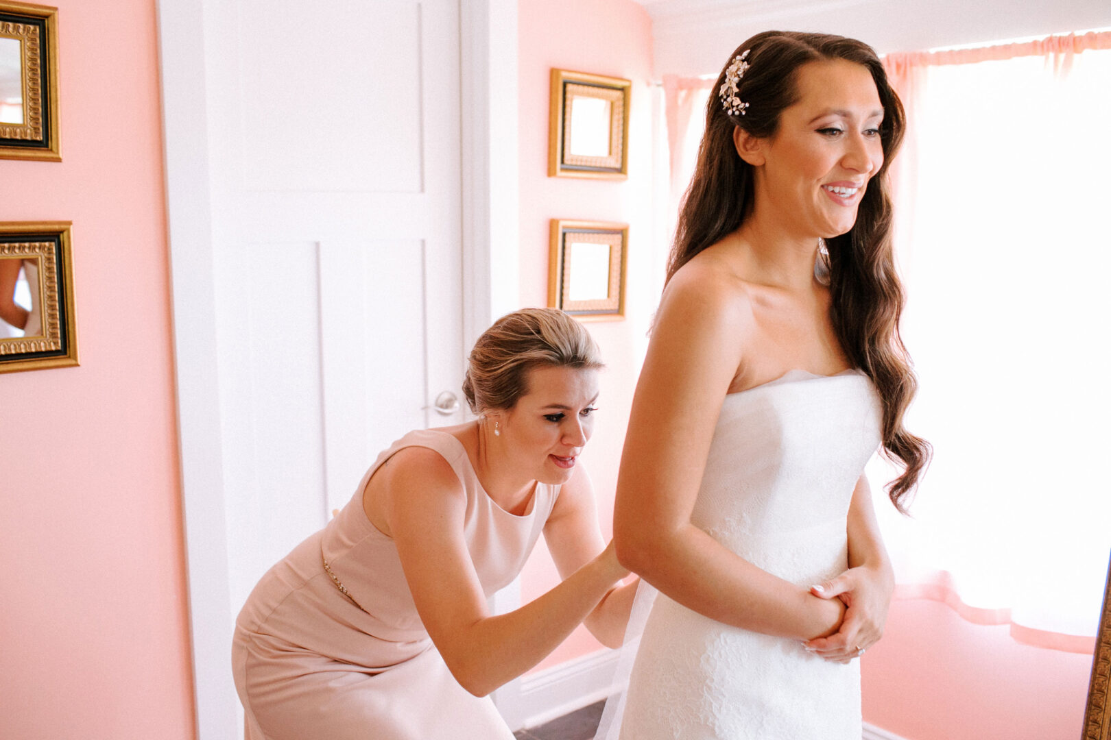 In a pink room, a woman assists a bride with perfecting her wedding dress, setting the stage for an unforgettable Willow Creek Winery wedding.