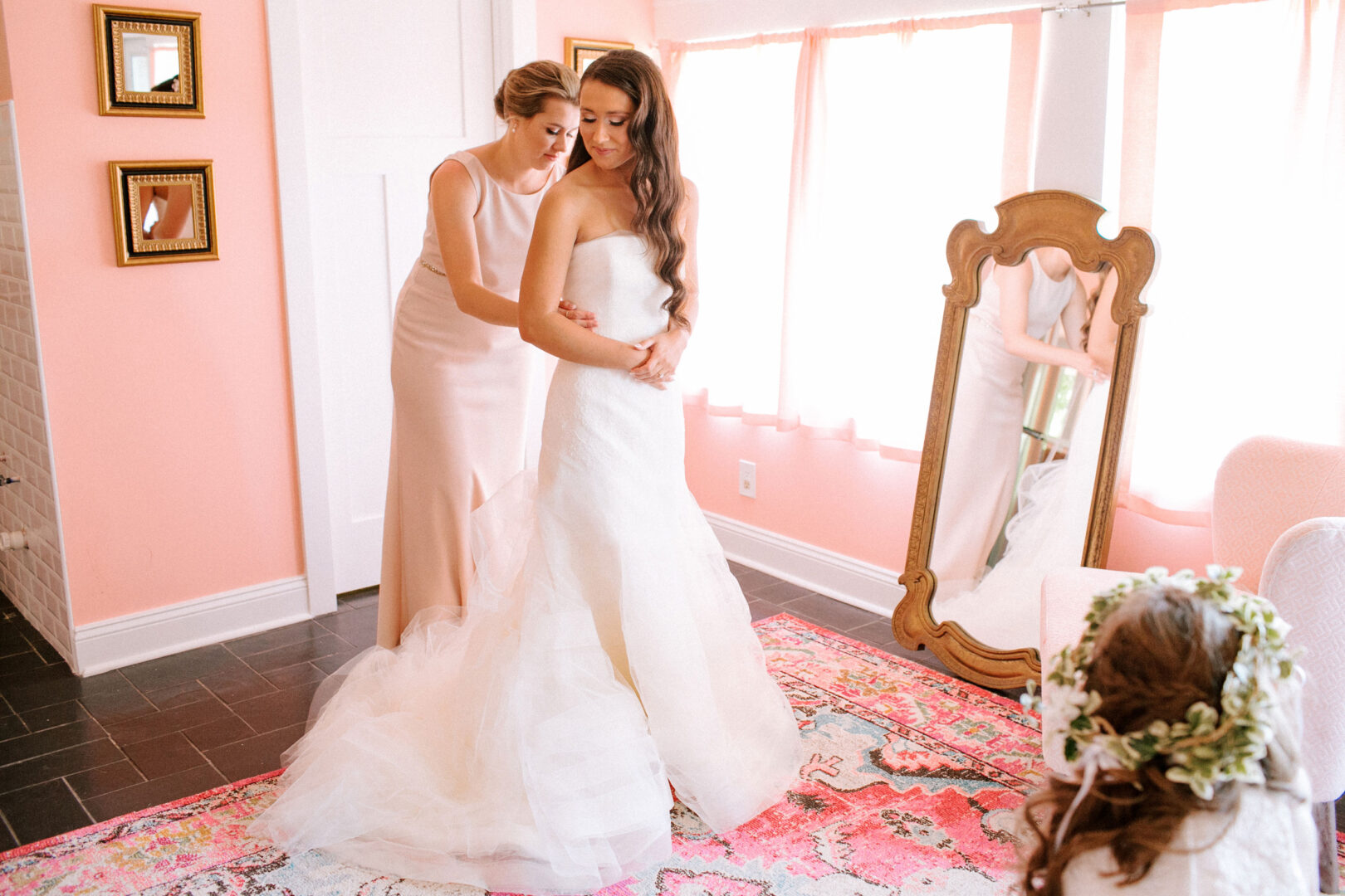 In a pink room adorned with a floral rug, a bride in a white gown is assisted by a woman in a blush dress. Nearby, at this Willow Creek Winery wedding, a child with a floral crown sits, their reflections captured in the full-length mirror.