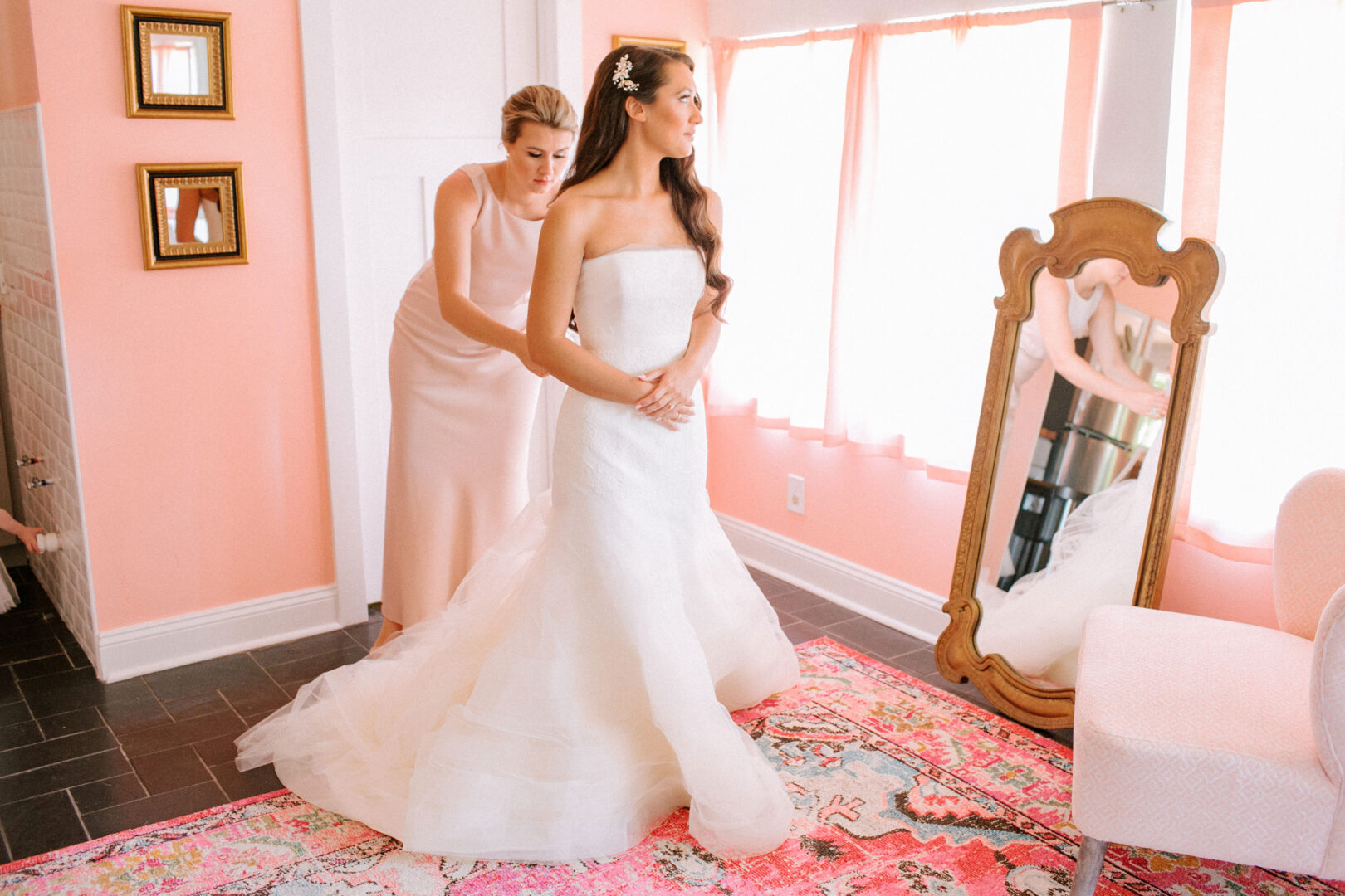 A woman in a white wedding dress is being assisted by another woman in a pink room, reminiscent of the charming ambiance of Willow Creek Winery Wedding, complete with a full-length mirror and a colorful rug.